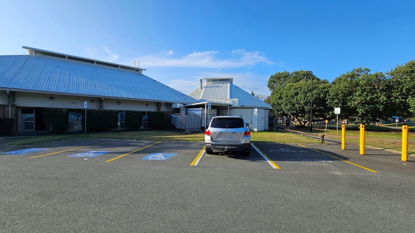 A car park with a silver SUV parked in a designated disabled parking space. The ground is marked with blue and white symbols. To the left, a large building with a corrugated metal roof and brick walls. On the right, yellow bollards line the edge of the parking area. In the background, there are trees and a grassy area with a path.