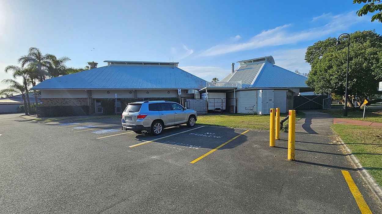 A car park with a silver SUV parked in a designated space on the left. The ground is asphalt with yellow parking lines. In the centre, a building with a metal roof and brick walls is visible. On the right, there are three yellow bollards and a pathway leading to a grassy area with a large tree. A lamppost and a sign are near the tree. Palm trees are in the background on the left.