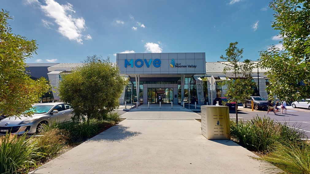 A two story building with a concrete and glass facade. bearing a sign reading "Moonee Valley" flanked by "Move" in larger letters is centered in the image. To the left, there is a grey sports car parked beside a small tree. To the right, pedestrians are walking towards the entrance. There is a yellow bin in the foreground on the right, and a series of short bushes line the pathway leading up to the building.