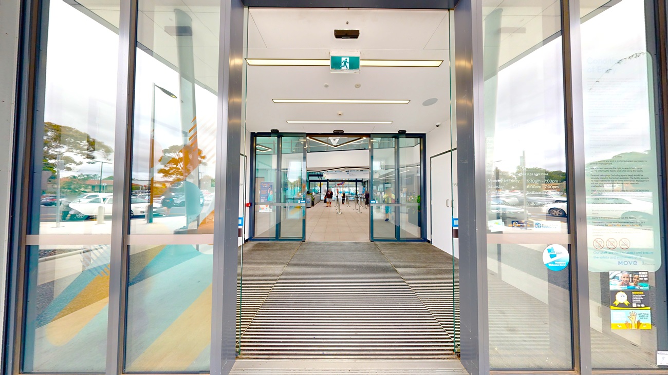 The facility entrance is outfitted with double automatic glass sliding doors, separated by a non-slippery mat.  Stickers and posters are affixed to the glass wall. The second door leads into the building's interior. White tiles and a white ceiling with stylish light fixtures adorn the interior. Visitors are also present.