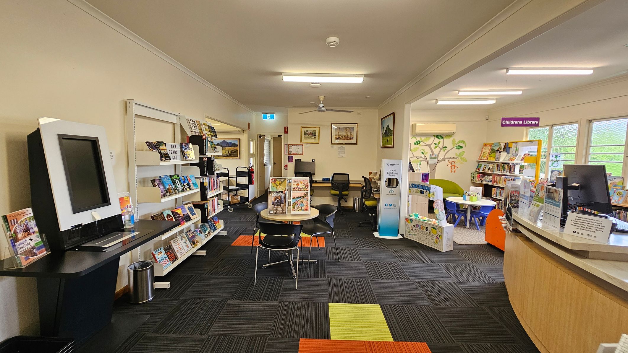 A library interior with a view through an open door. On the left, there are bookshelves filled with books and a poster on the wall. In the centre, a small round table with chairs is placed on a carpet with colourful square patterns. At the back, there are two computer stations with chairs. On the right, a glass partition displays more posters and a glimpse into another room with additional bookshelves and a counter. The ceiling has fluorescent lights.