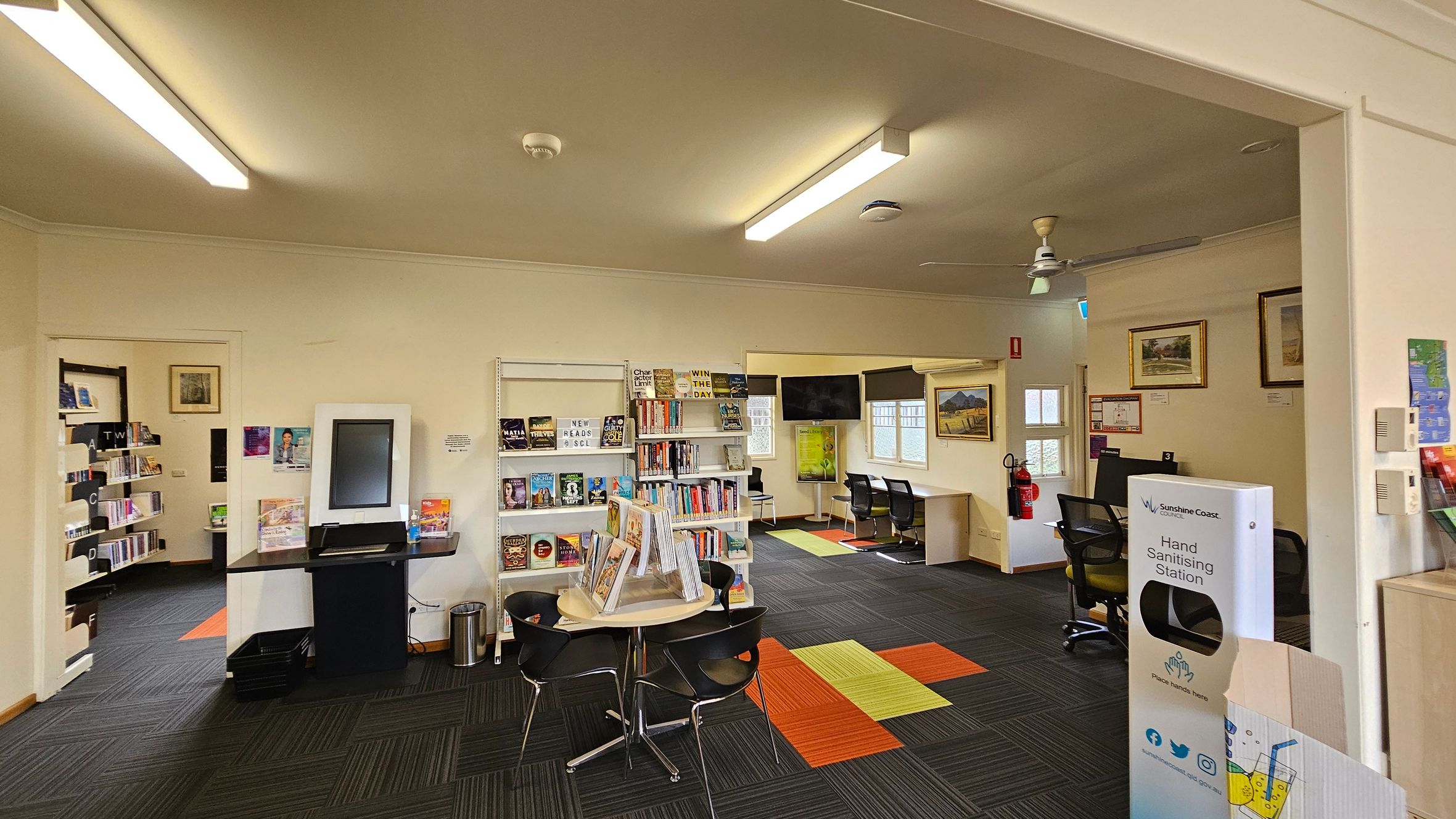 A library interior with a modern design. On the left, there are bookshelves filled with books and a digital kiosk. In the centre, a round table with chairs and a display of books. The right side features a hand sanitising station and framed artwork on the walls. The floor has a patterned carpet with green, orange, and black squares. The ceiling has fluorescent lights and a ceiling fan. In the background, there are windows and a small seating area with chairs and a table.