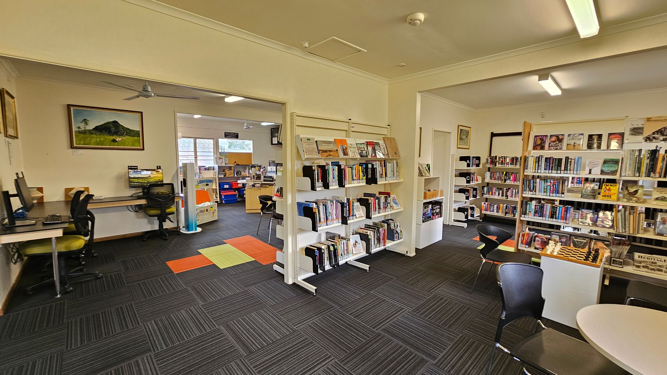 A library interior with bookshelves filled with books on the right. In the centre, a white shelving unit displays more books. On the left, there are computer workstations with chairs. A painting of a mountain hangs on the wall above the computers. The floor is covered with dark grey carpet tiles, accented with orange and green tiles. The ceiling has fluorescent lights and a ceiling fan. In the background, there is a window and more bookshelves.