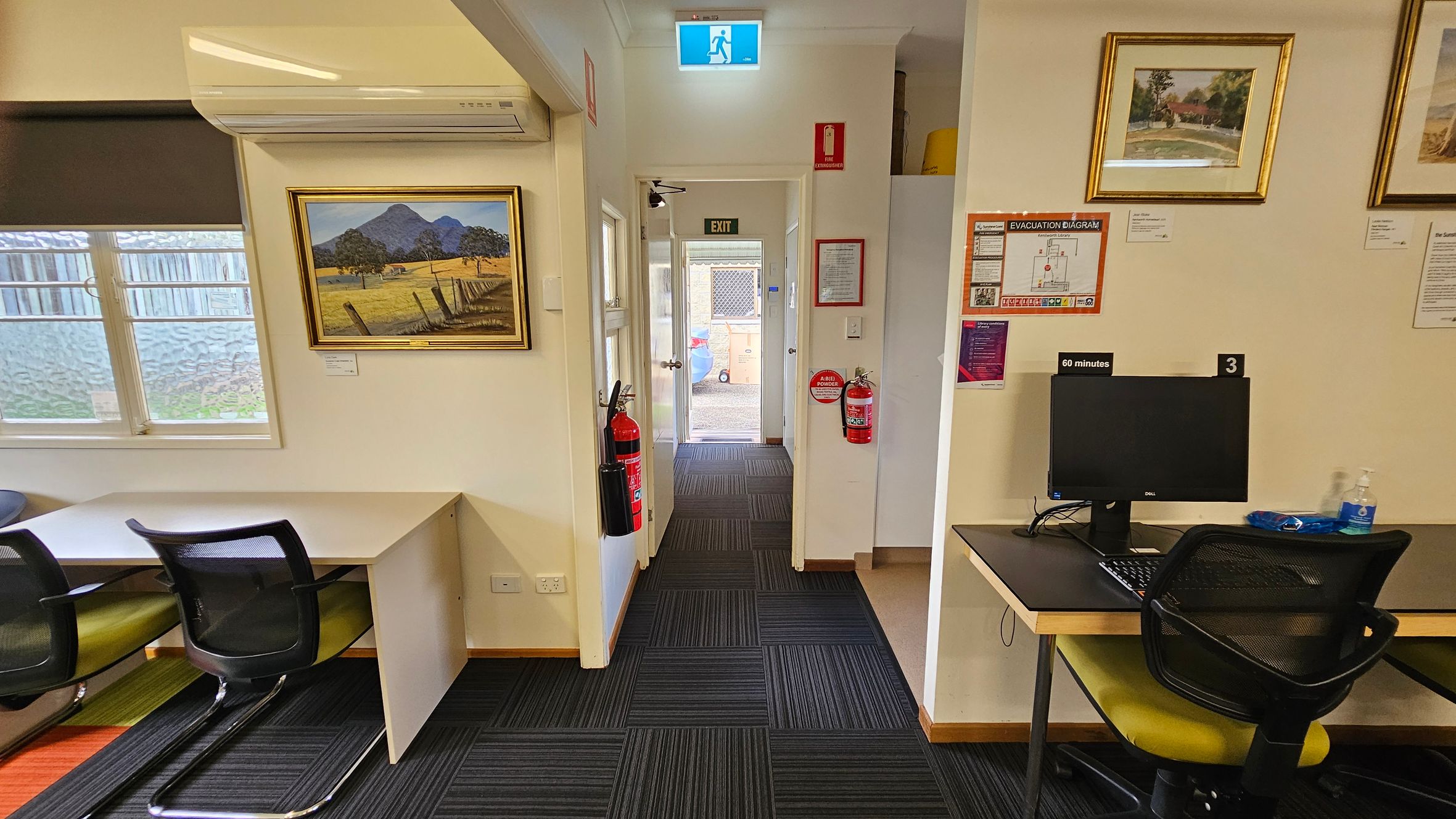 A room with a black and grey striped carpet. On the left, there's a window with a blind, a desk, and a chair. Above the desk is an air conditioning unit and a framed landscape painting. In the centre, a hallway leads to an exit door with a lit exit sign above. Fire extinguishers are mounted on the walls. On the right, there's a computer on a desk with a chair, and framed pictures on the wall. An evacuation diagram and other notices are displayed nearby.