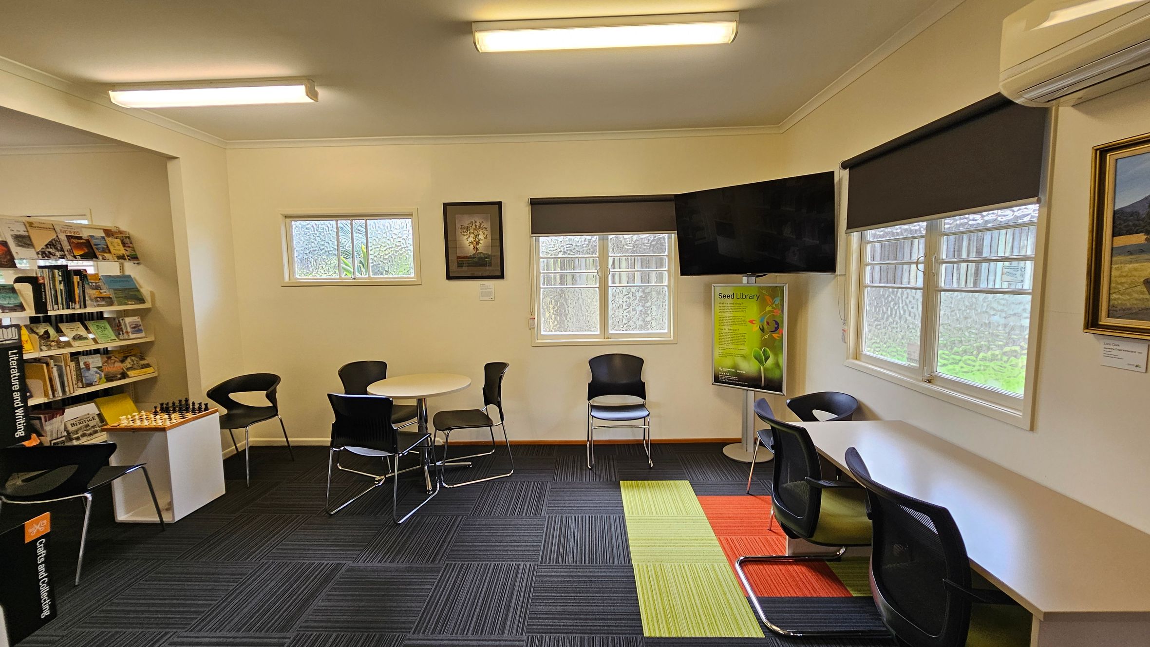A library room with a modern design. On the left, there are bookshelves filled with books and a chess set on a small table. In the centre, a round table with black chairs is placed near two small windows. A framed picture hangs between the windows. On the right, a long desk with chairs is positioned under another window. A large TV is mounted above a poster stand. The floor has a mix of dark, green, and orange carpet tiles, and the ceiling has fluorescent lights.