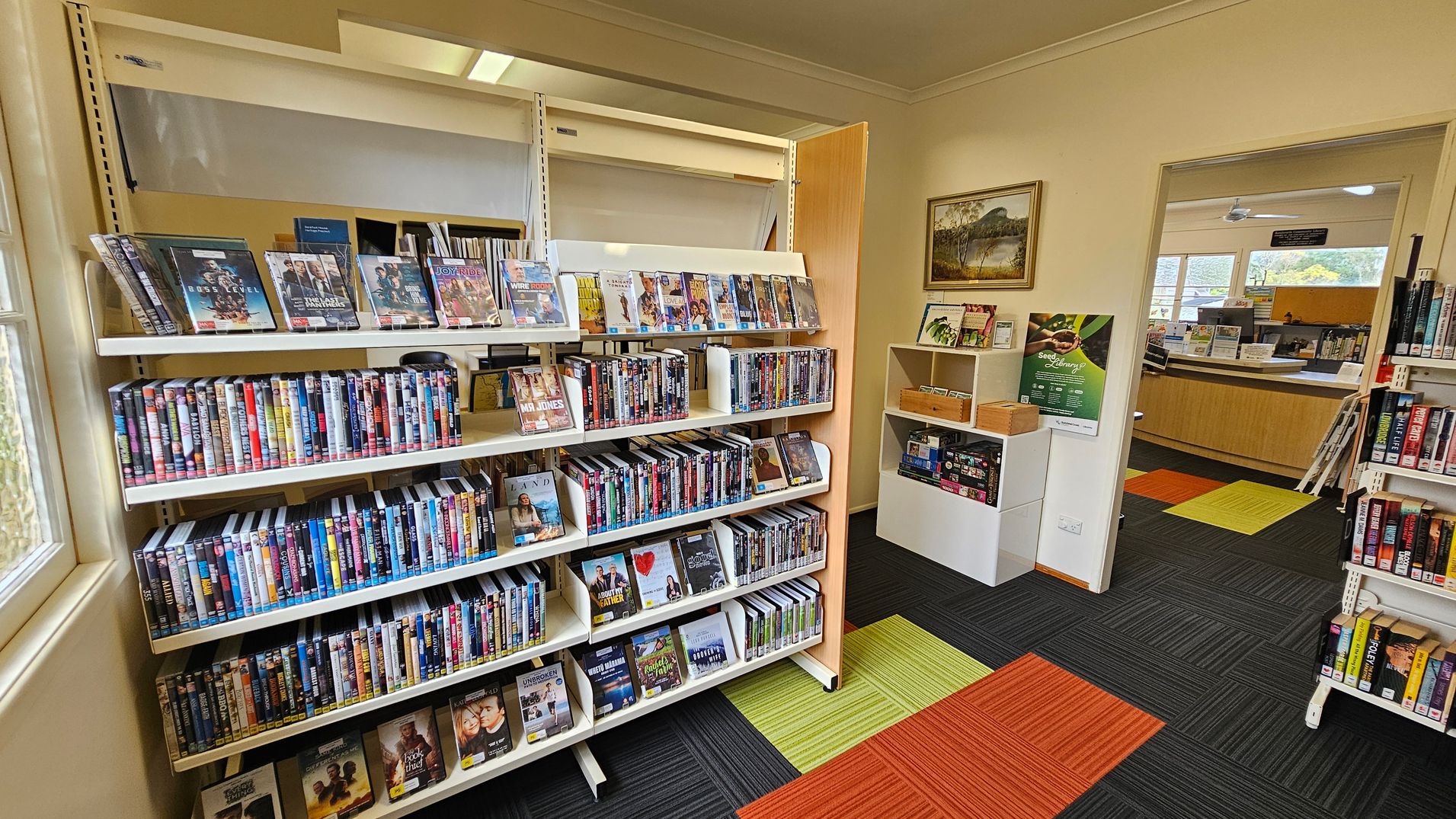 A library interior with shelves filled with DVDs on the left. The DVDs are arranged in rows, with some displayed facing forward. To the right, there's a small white shelf with brochures and a poster on the wall above it. An open doorway leads to another room with a wooden counter and large windows. The floor is covered with carpet tiles in black, green, and orange. A framed landscape painting hangs on the wall near the doorway.