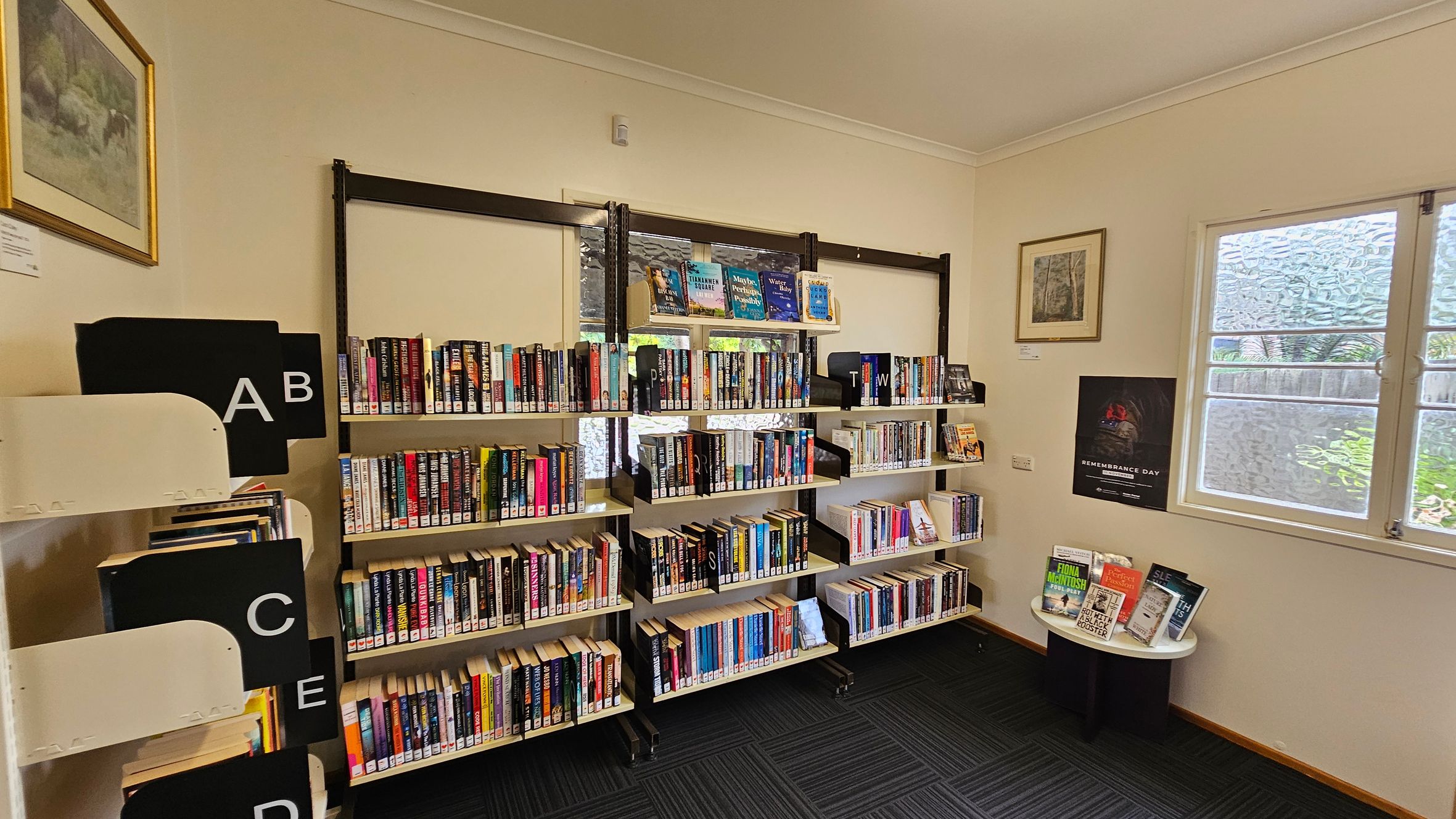 Bookshelves filled with books are against a cream-coloured wall. On the left, there are labelled sections with letters A to E. In the middle, three shelves hold books with dividers marked with letters. On the right, a small round table displays more books. Above it, a framed picture and a Remembrance Day poster are on the wall. A window with frosted glass is next to the table. The floor is carpeted with a dark pattern.