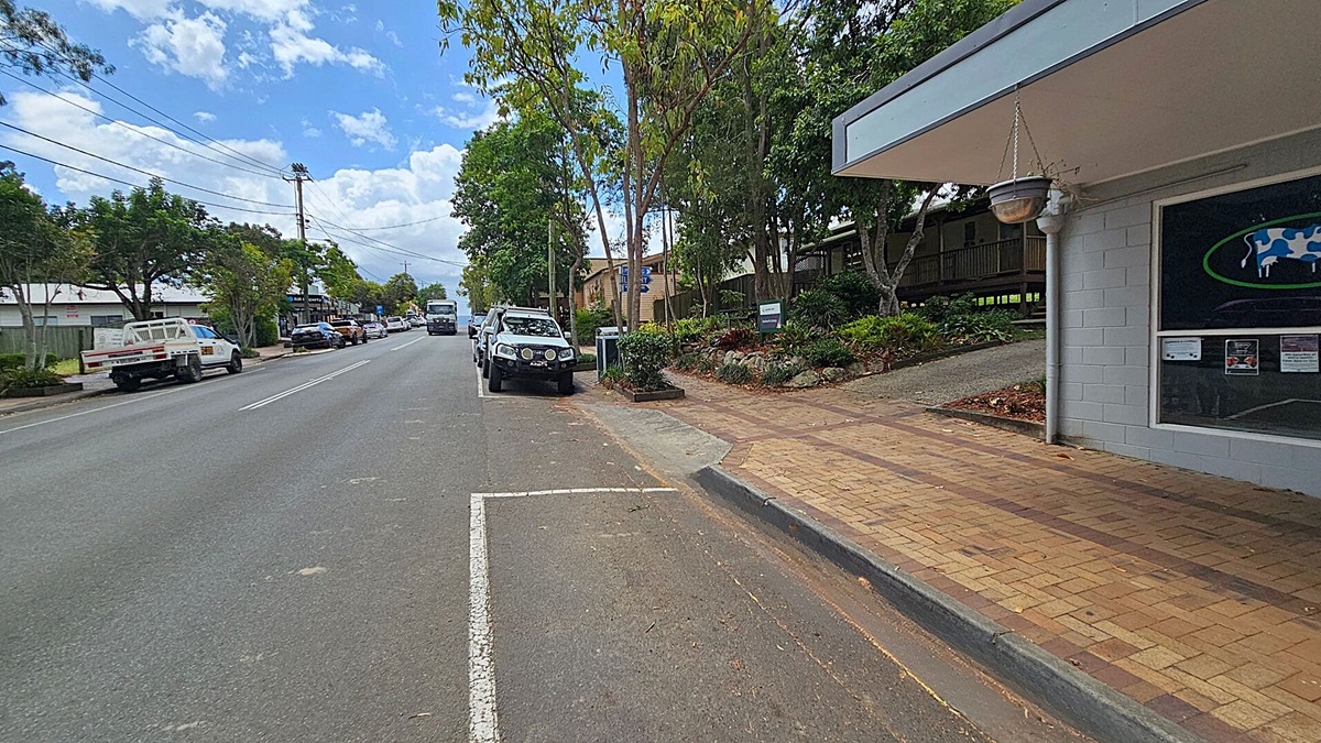 A street scene with a paved footpath on the right, bordered by a grey building with a hanging pot and a window displaying a cow graphic. Several cars are parked along the road, including a white vehicle in the foreground. Trees line the street, providing shade. On the left, more vehicles are parked, and a few buildings are visible in the distance. The road is marked with white lines, and the sky is partly cloudy.