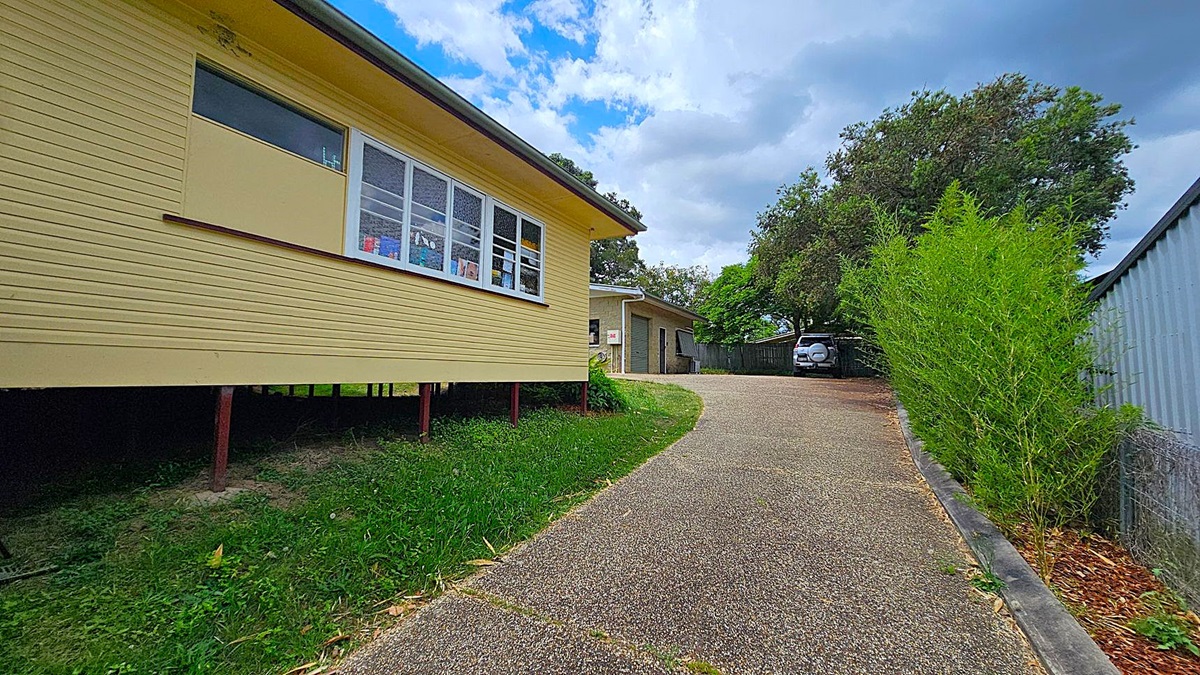 A yellow house on the left with large windows, elevated on stilts. A gravel driveway curves to the right, leading to a garage and a parked car. On the right, a tall green hedge runs alongside a metal fence. Trees are visible in the background.