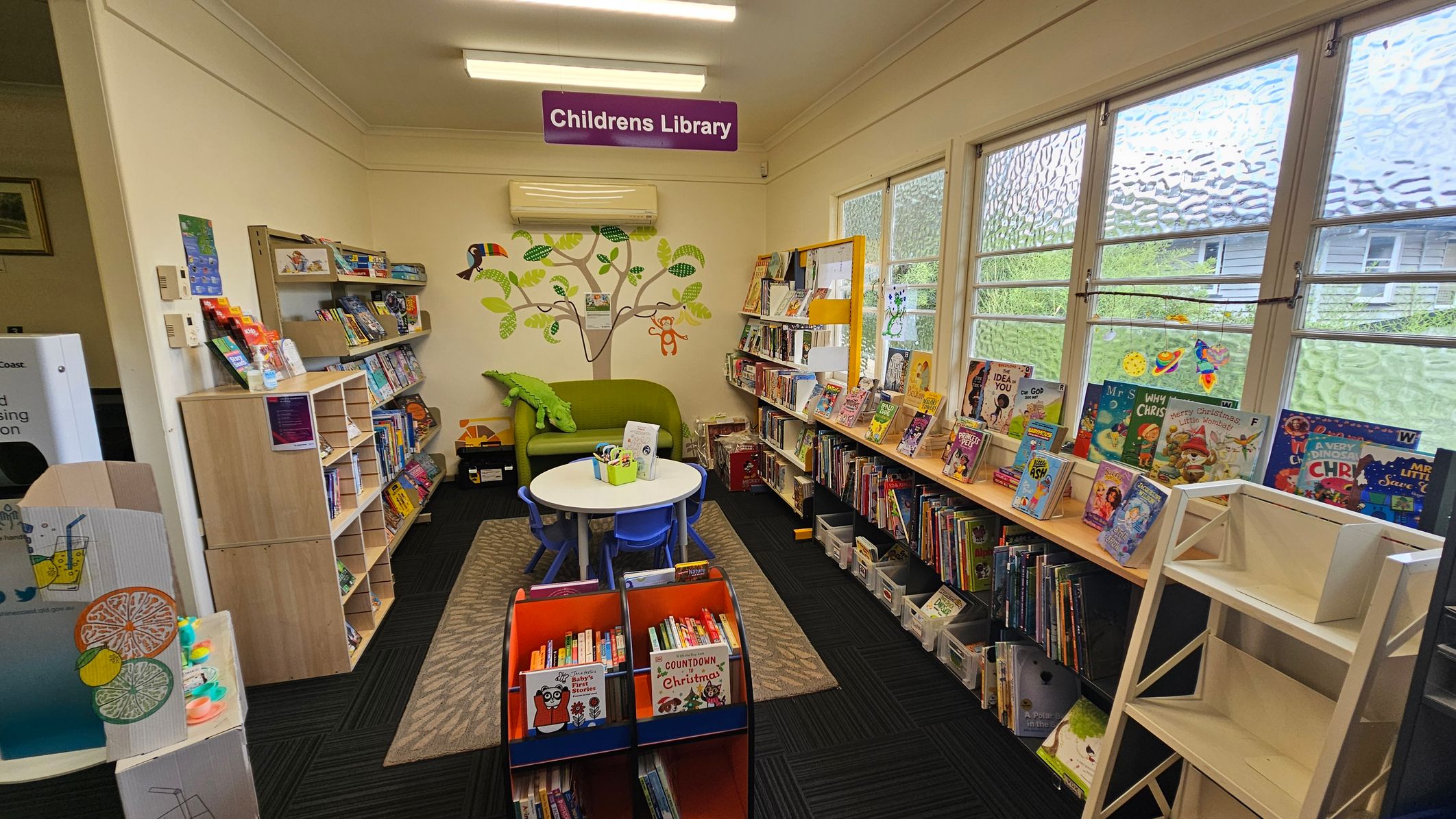 A library interior with a children's section on the left, featuring bookshelves filled with colourful books and a small play area with a lemonade stand. In the centre, a curved wooden counter displays brochures and a "Reservations" sign. Behind the counter, a computer and office supplies are visible. The room has fluorescent lighting and a ceiling fan. A wall clock is mounted on the right wall, and large windows provide natural light. The floor is carpeted with a dark pattern.
