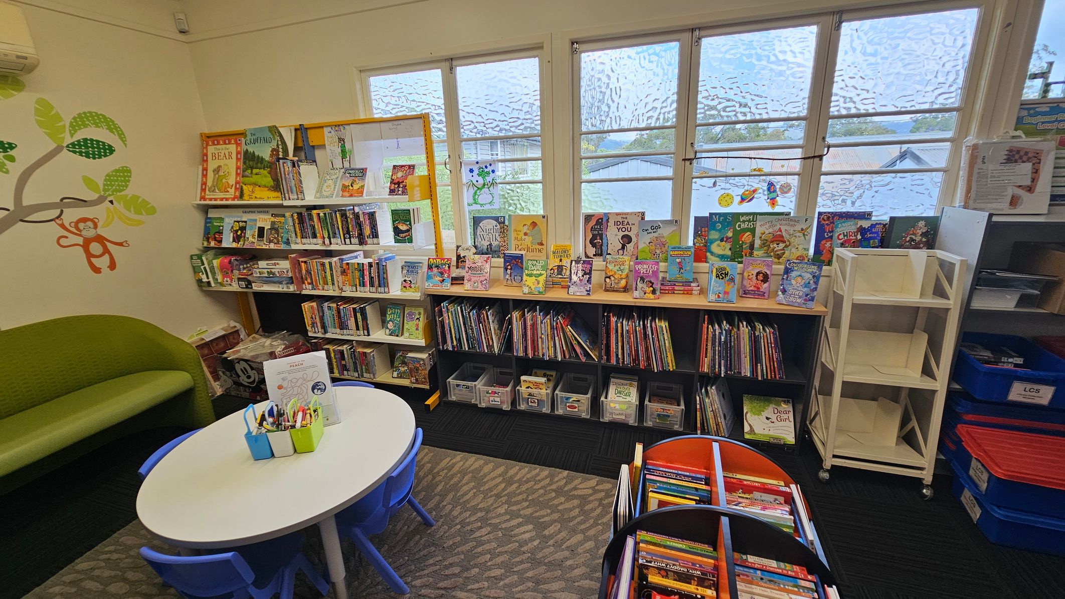 A cosy children's reading area with a green sofa on the left, decorated with a wall mural of a tree and a monkey. In the centre, a small round table with blue chairs is set up for activities, holding containers with pens. Bookshelves line the walls, filled with colourful children's books. Large windows in the background let in natural light. On the right, there are empty white shelves and blue storage bins. The floor is carpeted with a patterned rug.