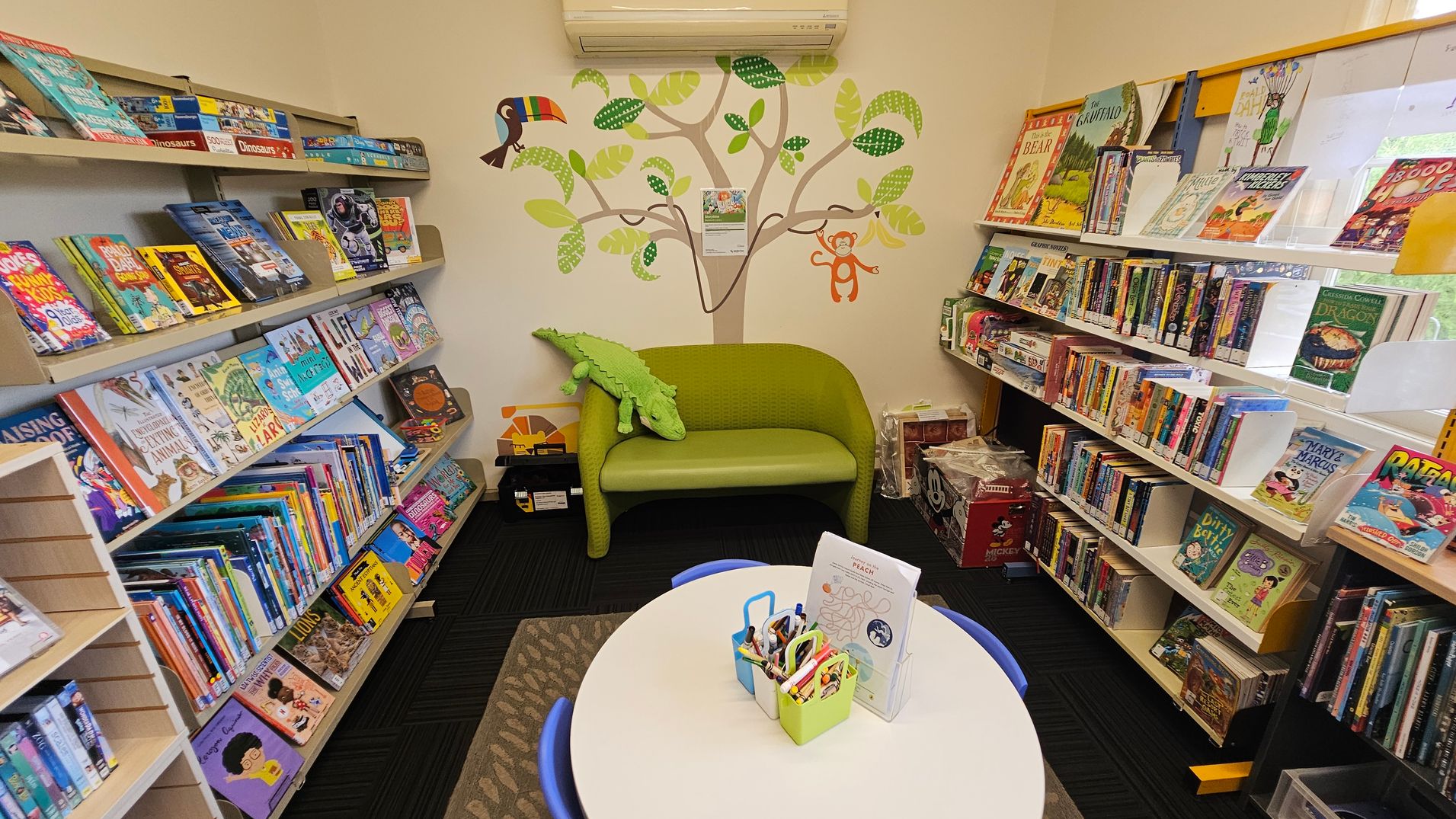 A cosy children's reading area with bookshelves on both sides filled with colourful books. In the centre, a small green sofa with a plush crocodile toy sits against a wall decorated with a tree mural featuring a toucan and a monkey. In the foreground, a round table with blue chairs holds containers of crayons and paper. The floor is carpeted in dark tones, and the ceiling is not visible.