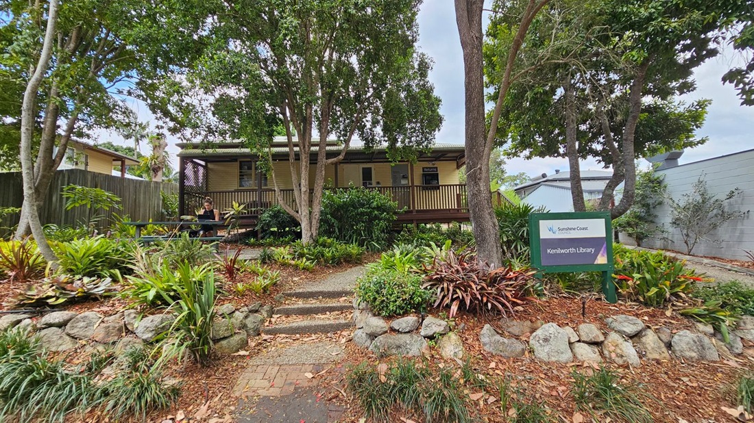A small building with a wooden deck is surrounded by lush greenery and trees. A person sits at a picnic table on the left, using a laptop. A sign on the right reads "Kenilworth Library" with "Sunshine Coast" branding. The foreground features a stone-bordered garden with various plants and a pathway leading up to the building. A wooden fence is visible on the left, and a grey wall is on the right.