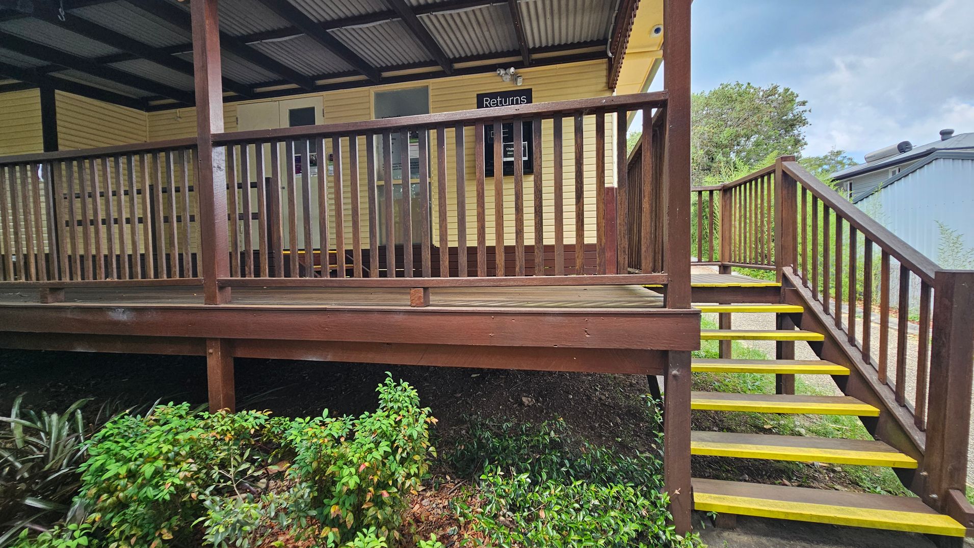 A wooden deck with a railing and a set of stairs leading up to it. The stairs have yellow edges for visibility. The building is painted yellow with a sign that reads "Returns" above a window. The roof is corrugated metal. In the foreground, there are green shrubs and plants. To the right, a path leads away from the stairs, and a grey building is partially visible in the background.