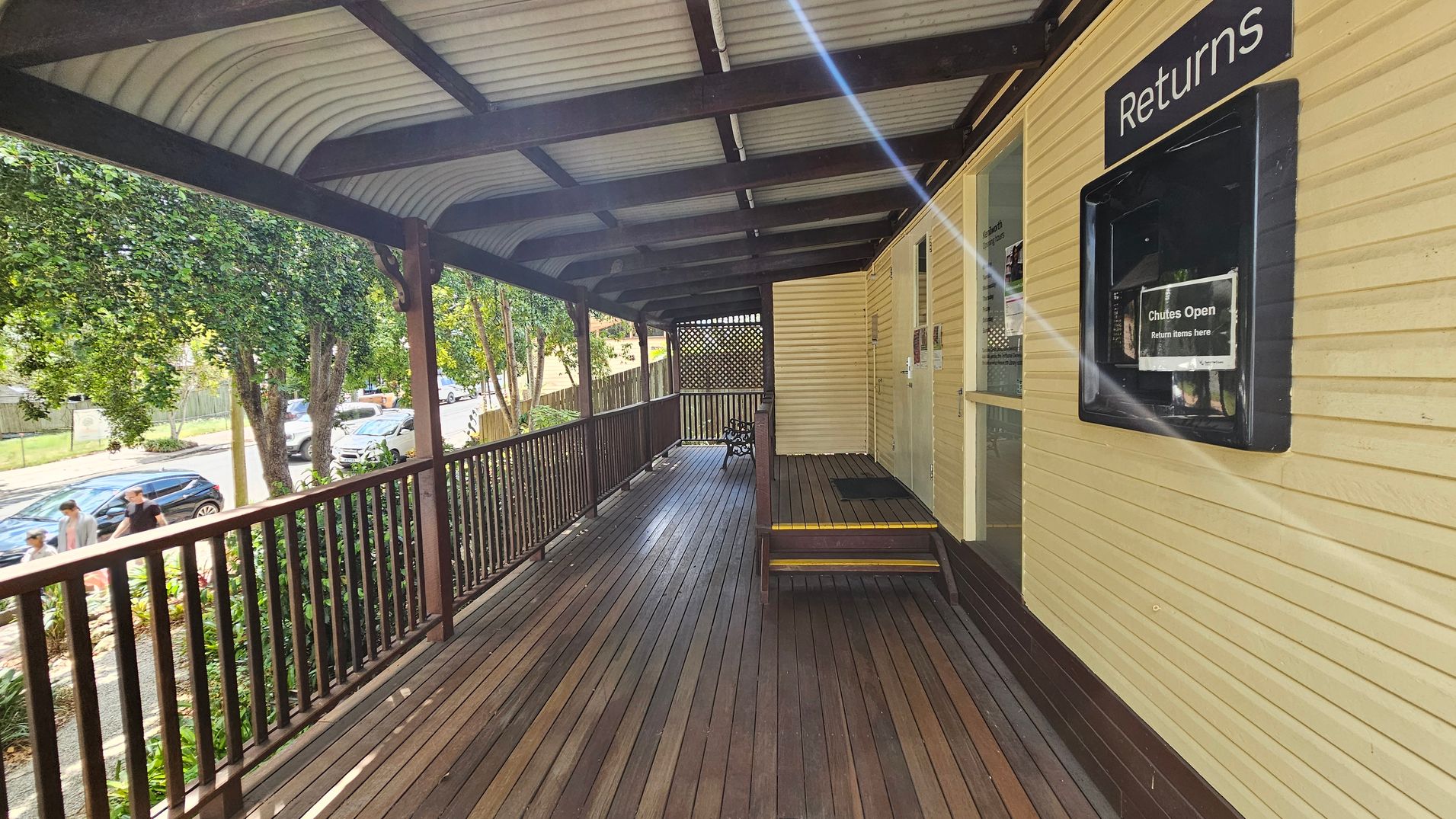 A wooden deck with a corrugated metal roof. On the right, a yellow wall features a "Returns" slot with a sign indicating "Chutes Open." The deck has a wooden railing, and a small set of steps leads to a door. On the left, trees and parked cars line the street, with a few people walking on the pavement. The floor is made of wooden planks.