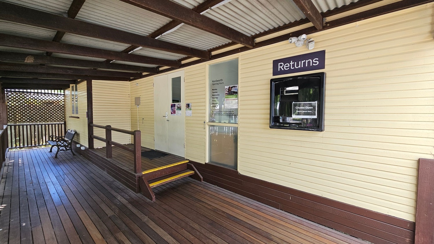 A wooden deck with a bench on the left side, leading to a building with cream-coloured walls. The building has a "Returns" slot on the right, and a glass door with opening hours listed. A small set of steps with yellow edges leads up to the door. The ceiling is made of corrugated metal with wooden beams. A lattice screen is visible in the background on the left.