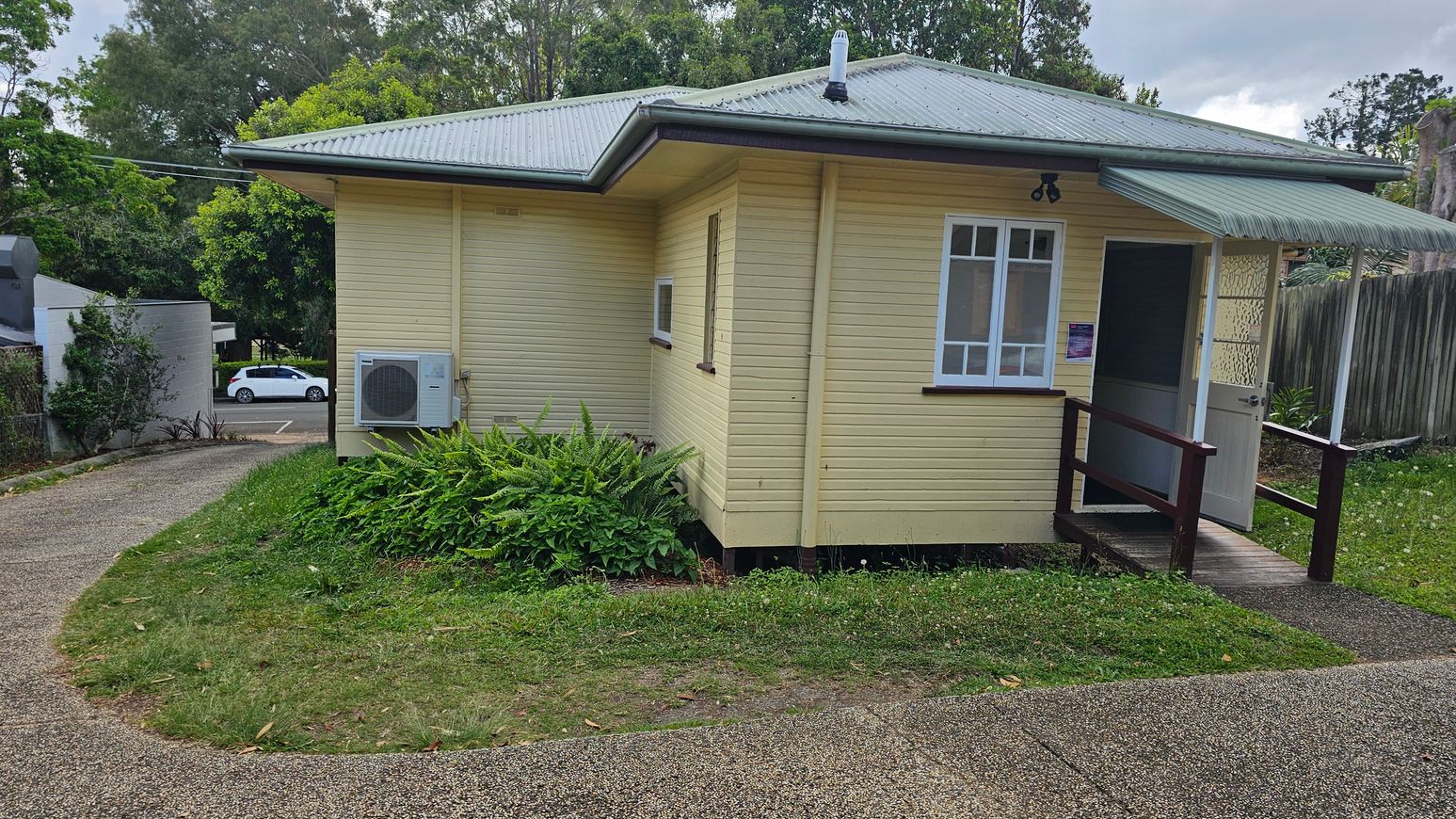 A small, single-storey yellow house with a grey metal roof. On the right, there's a partially open door with a small awning above it. A window is next to the door. An air conditioning unit is mounted on the left side of the house. The house is surrounded by grass and ferns. A paved path curves around the house, leading to a road where a white car is parked. Trees and a wooden fence are in the background.