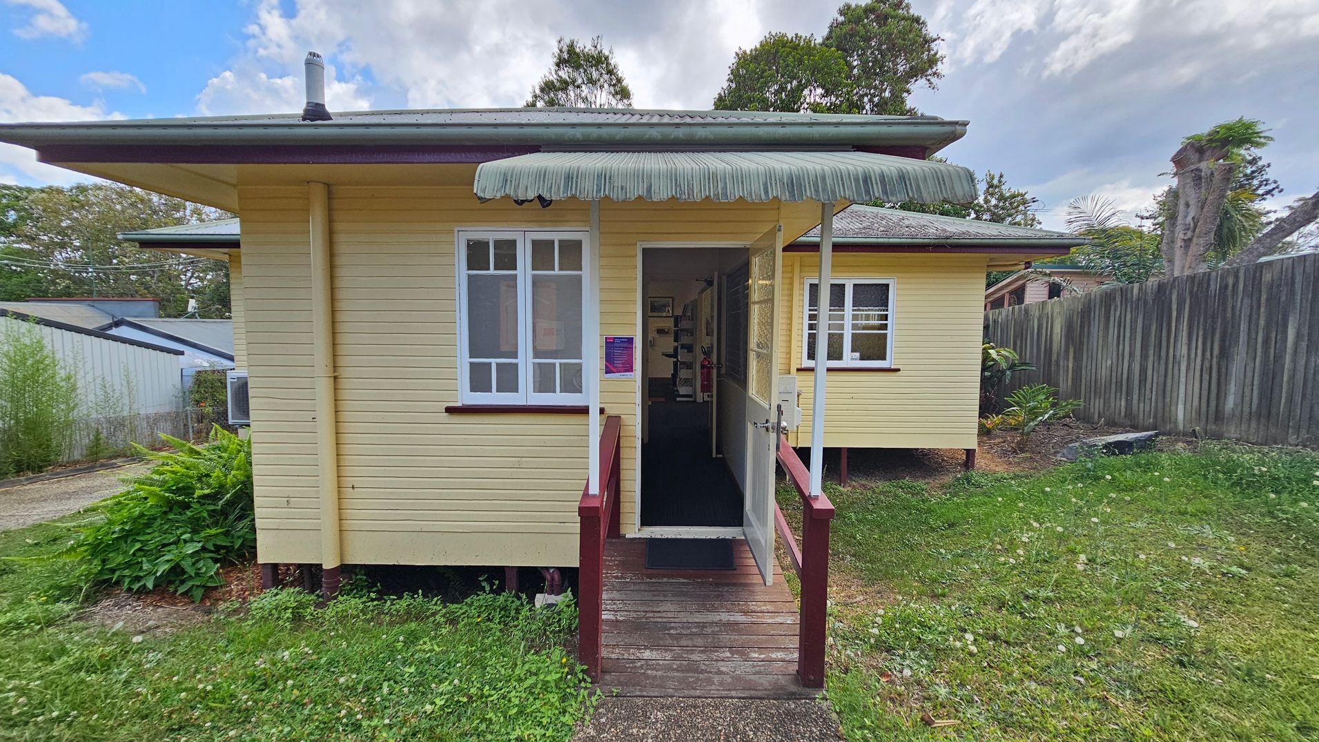 A small, single-storey yellow wooden house with a green corrugated awning above the entrance. The entrance has a wooden ramp with red railings leading to an open door. Two white-framed windows are visible on either side of the door. The house is elevated slightly on stilts. To the right, there's a wooden fence and some greenery. The left side shows a neighbouring building and more plants. The grass in the foreground is patchy with some weeds.