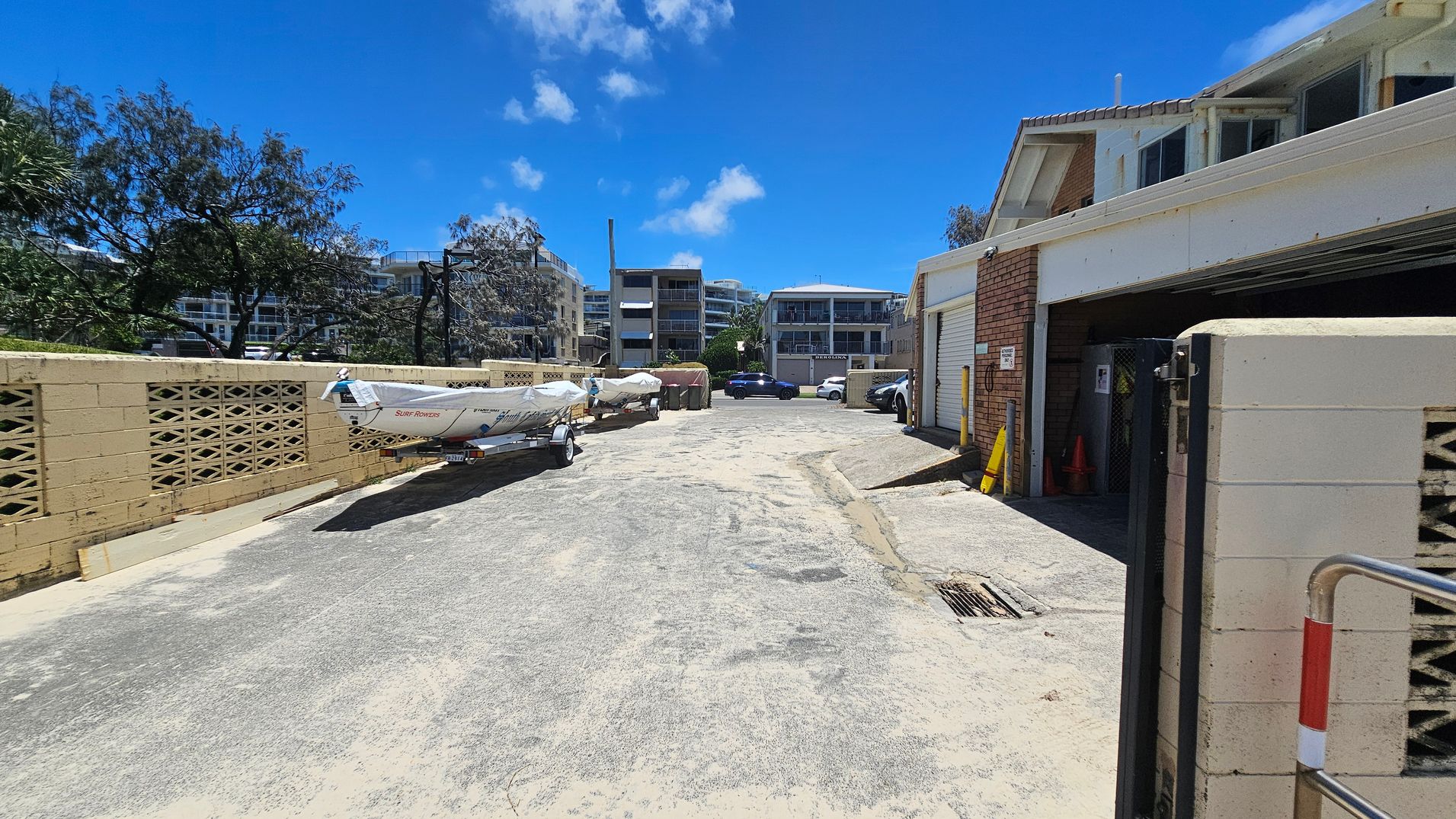 A narrow alleyway with a concrete surface. On the left, there's a beige brick wall with a boat on a trailer parked alongside it. Trees are visible behind the wall. On the right, a building with a garage door and a partially open gate. In the background, several modern apartment buildings and parked cars.