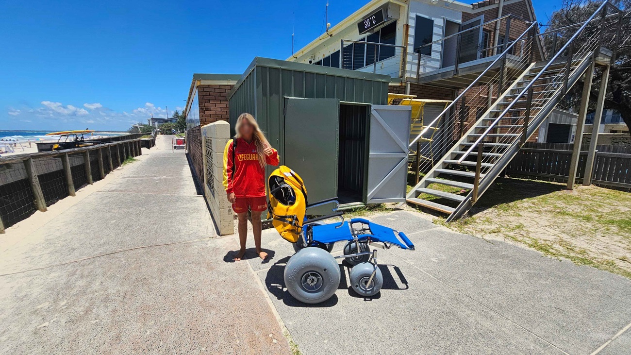 A person in a red and yellow lifeguard uniform stands on a paved path next to a blue beach wheelchair with large grey wheels. Behind them is a green metal shed with open doors. To the right, a staircase leads up to a building with a balcony. On the left, the path runs alongside a sandy beach with a yellow lifeguard boat visible in the distance.