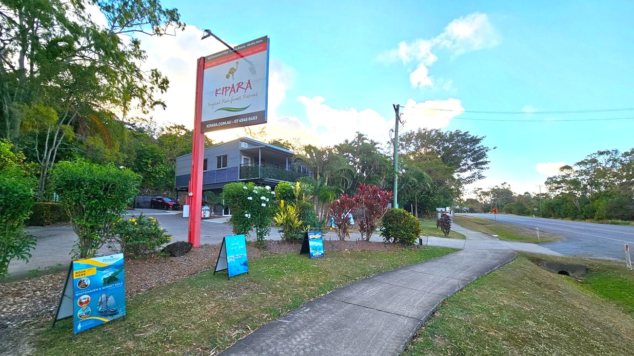 A paved footpath runs alongside a road on the right, bordered by grass. On the left, a large sign reads "Kipara Tropical Rainforest Retreat" with additional promotional signs below it. Behind the signs, a building is partially visible, surrounded by lush greenery and various plants. The area is well-maintained, with a mix of trees and shrubs. The sky is clear with some clouds.