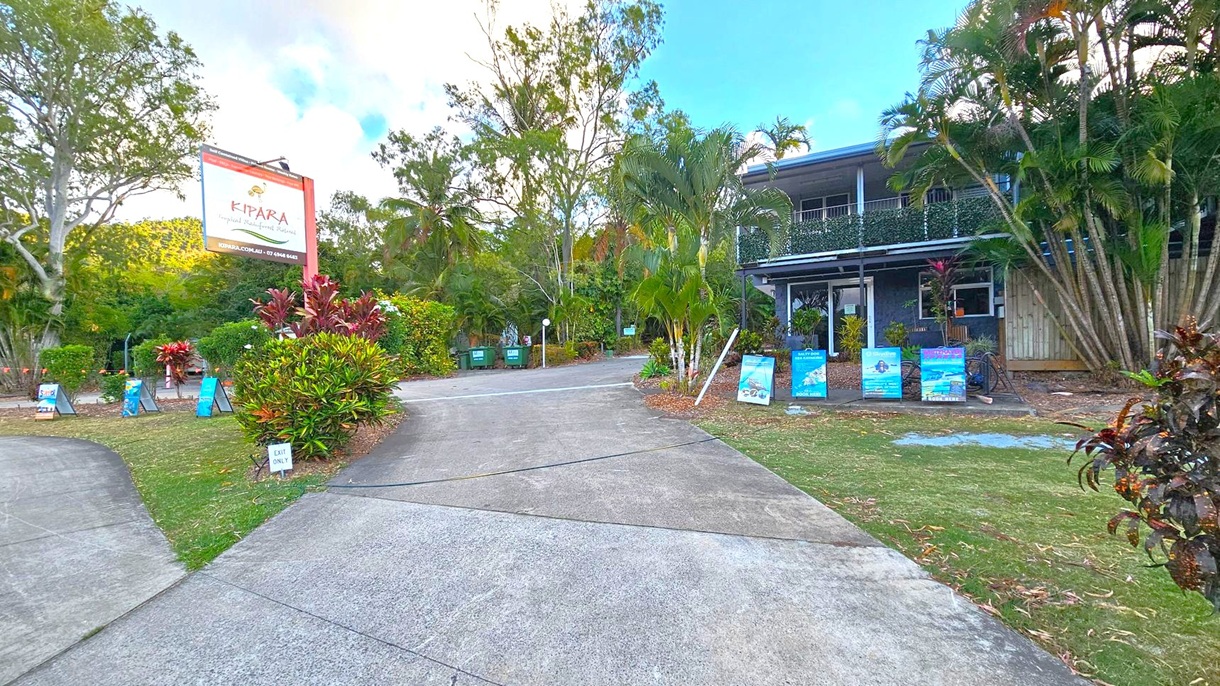A driveway leads to a building on the right, partially obscured by lush greenery and palm trees. Several signs and posters are displayed along the path. On the left, a large sign reads "Kipara Tropical Rainforest Retreat" with contact information. The area is surrounded by dense foliage, including trees and shrubs. The ground is a mix of concrete and grass, with a small "Exit Only" sign visible. The sky is partly cloudy.