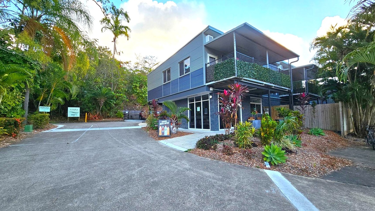 A modern two-storey building with a metal exterior and a balcony covered in greenery is on the right. The ground floor has large glass windows and a signboard outside. The driveway leads up to the building, with tropical plants and trees lining both sides. On the left, there are signs indicating parking areas. The background is filled with dense foliage, and the sky is partly cloudy. The ground is paved with a mix of concrete and asphalt.