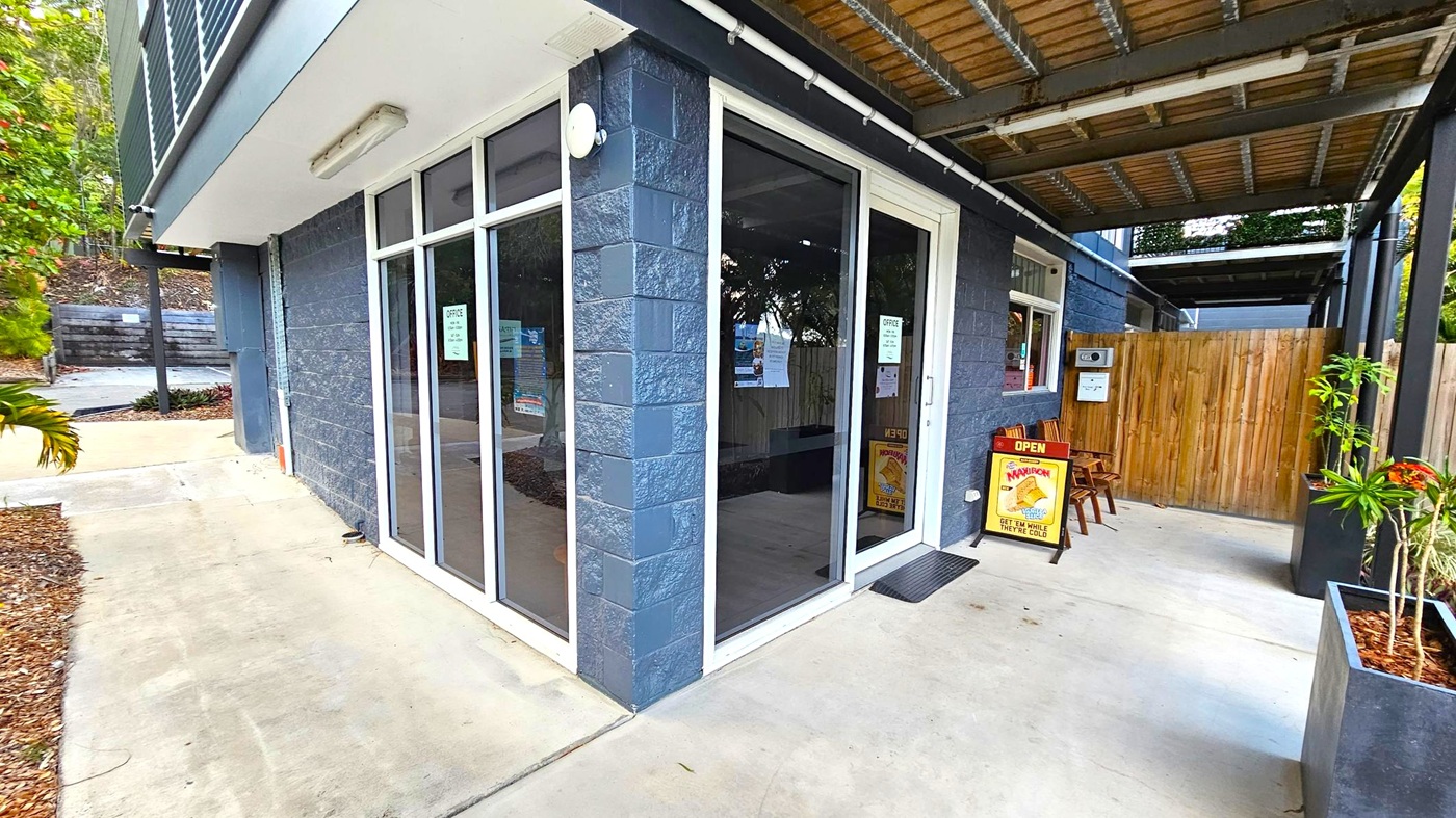 A building corner with large glass windows and a door displaying signs. The exterior walls are made of dark grey bricks. A wooden fence is visible on the right, with a small seating area featuring two chairs and a signboard. A potted plant is placed nearby. The ceiling has exposed beams and a light fixture. The ground is concrete, with a small garden area on the left containing mulch and plants.