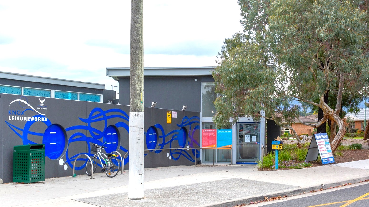 Knox Leisureworks is a low-set building with grey-painted walls. The brand name and logo can be seen on the outside walls of the venue. A bicycle parking rack is accessible at the concrete pathway just before the main entrance door of the facility.