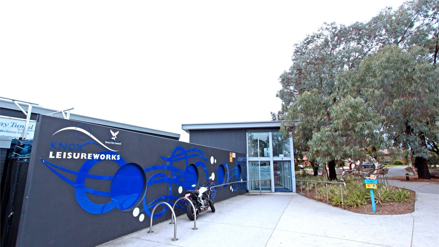 Motorcycles can also be parked in the accessible parking spaces in front of the facility. A number of trees with dense foliage can be found in front of the Knox Leisureworks building, as well as a landscape with ornamental shrubs. Half of the two-storey entrance building is made of glass walls.