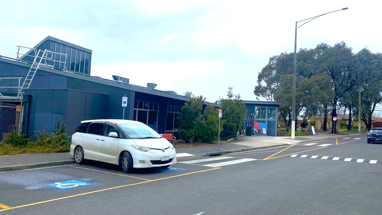 A white car is parked in one of the accessible parking spaces beside the sidewalk and directly opposite the main entry point of Knox Leisureworks. At the individual parking spaces is an accessible parking sign for persons with disabilities.