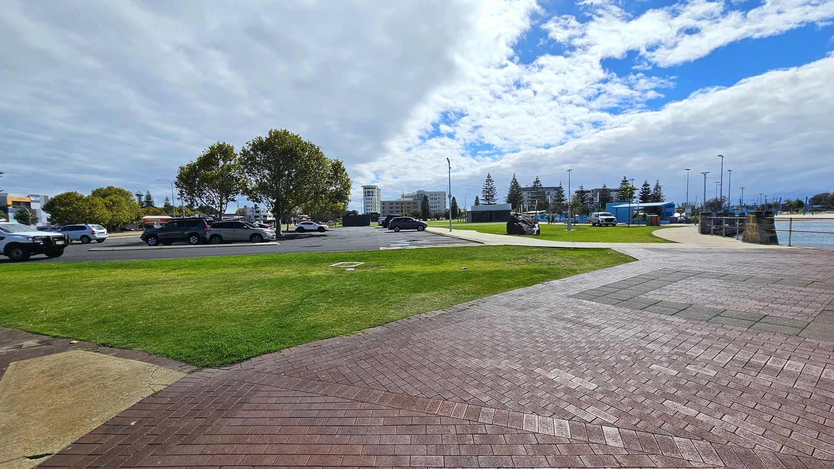 A paved area with a mix of red and grey bricks is in the foreground, leading to a grassy section. To the left, several cars are parked near a row of trees. In the background, there are buildings and more parked cars. On the right, a pathway curves towards a waterfront area with a low stone wall and a body of water. The sky is partly cloudy with patches of blue visible.