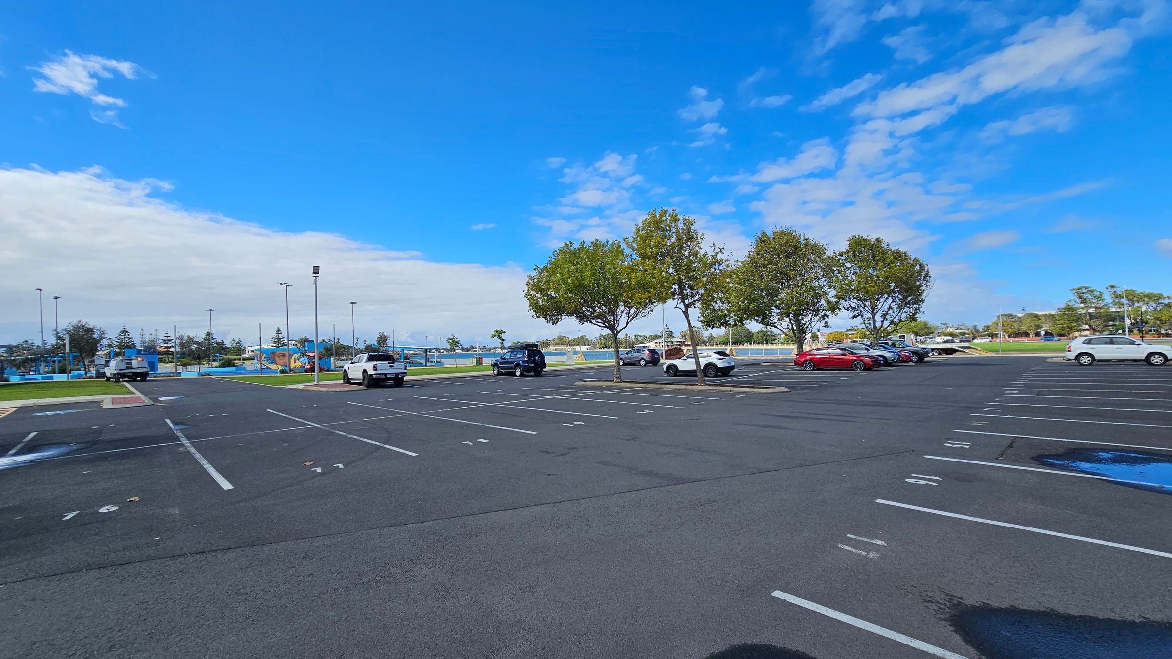 An expansive car park with marked parking spaces, some occupied by vehicles. On the right, several cars are parked near a row of trees. In the middle, a few more cars are scattered across the lot. The left side shows a grassy area with a few trees and a distant view of a sports field or park. The ground is asphalt with visible parking lines, and there are a few puddles. The background features more trees and some structures, possibly part of a recreational area.