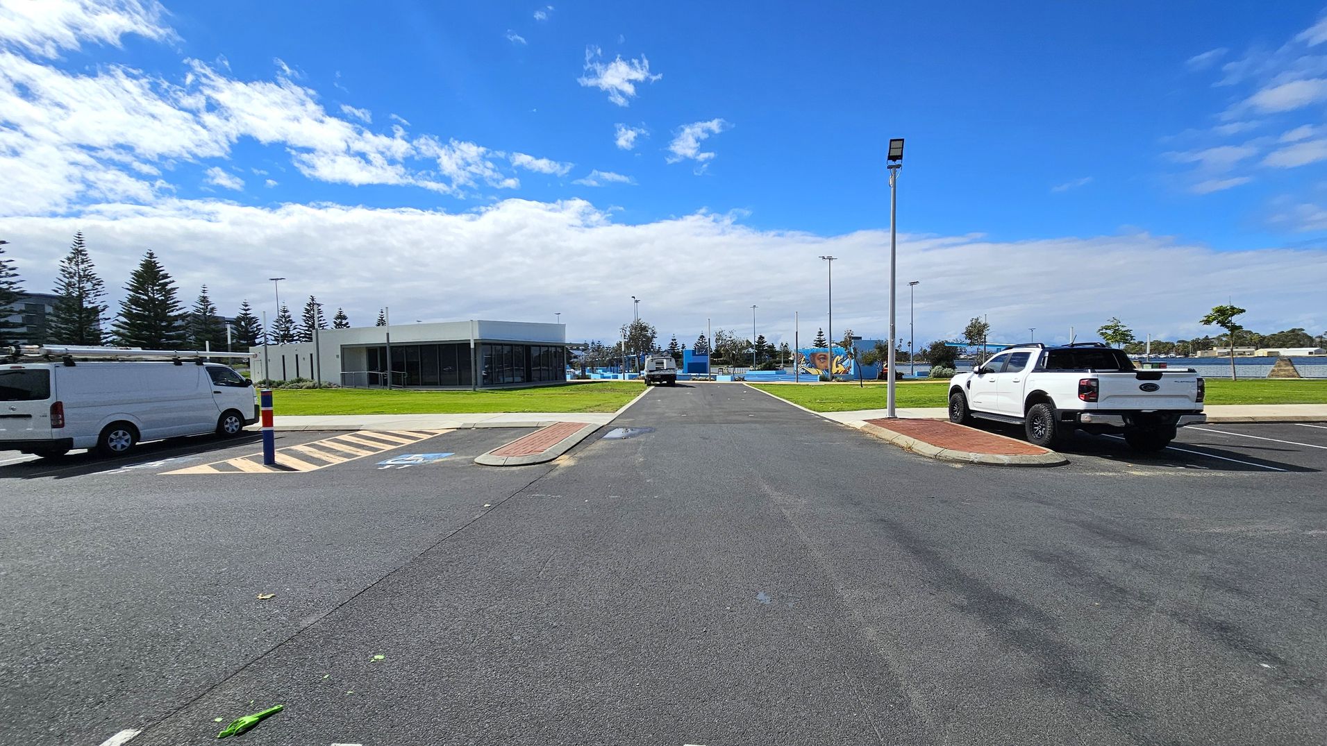 A car park with a white van on the left and a white pickup truck on the right. In the centre, a road leads to a building with large windows and a flat roof. The background features a grassy area with scattered trees and a playground. A tall streetlight is on the right side of the road. The sky is partly cloudy.