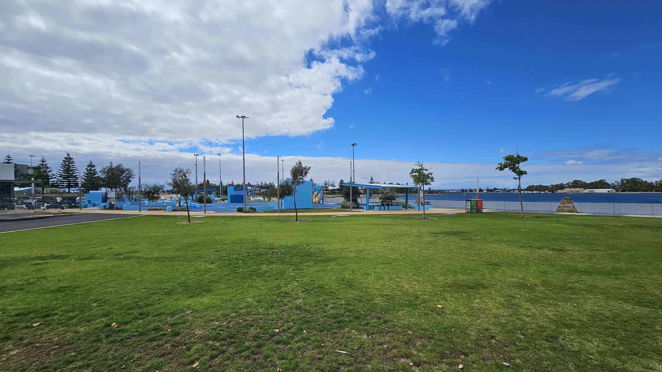 A grassy park area with patches of green and brown in the foreground. In the middle, there are several small trees and a pathway leading to a blue-painted recreational area with structures and poles. To the right, a body of water is visible, bordered by a fence. The background features more trees and a partly cloudy sky.