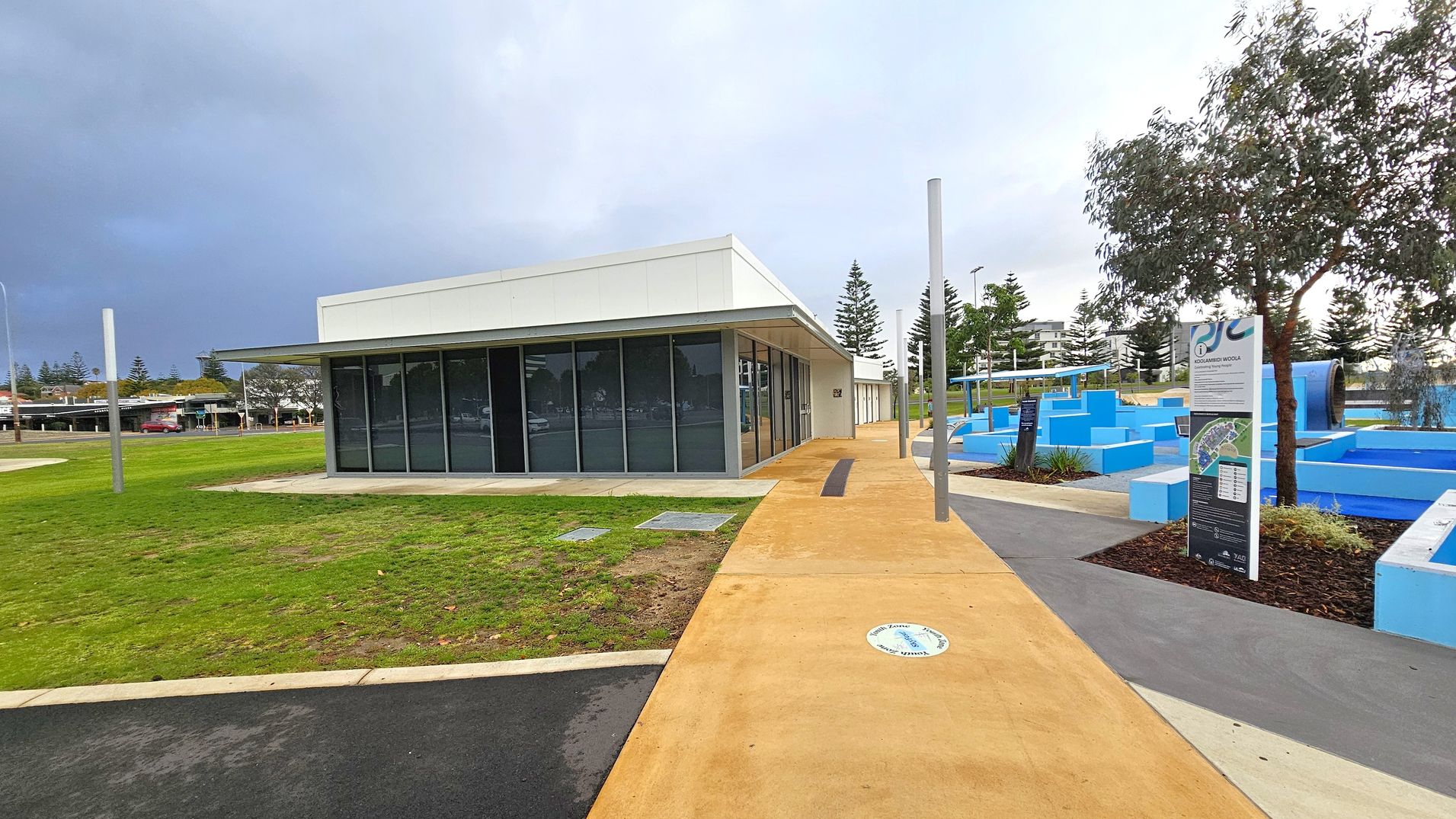 A modern, single-storey building with large glass windows on the left, surrounded by a grassy area. A paved path leads to the building, with a circular sign on the ground. On the right, there are blue geometric structures and a sign with information. A tree is nearby, and several tall poles are scattered around. In the background, there are more trees and a few buildings. The sky is overcast.