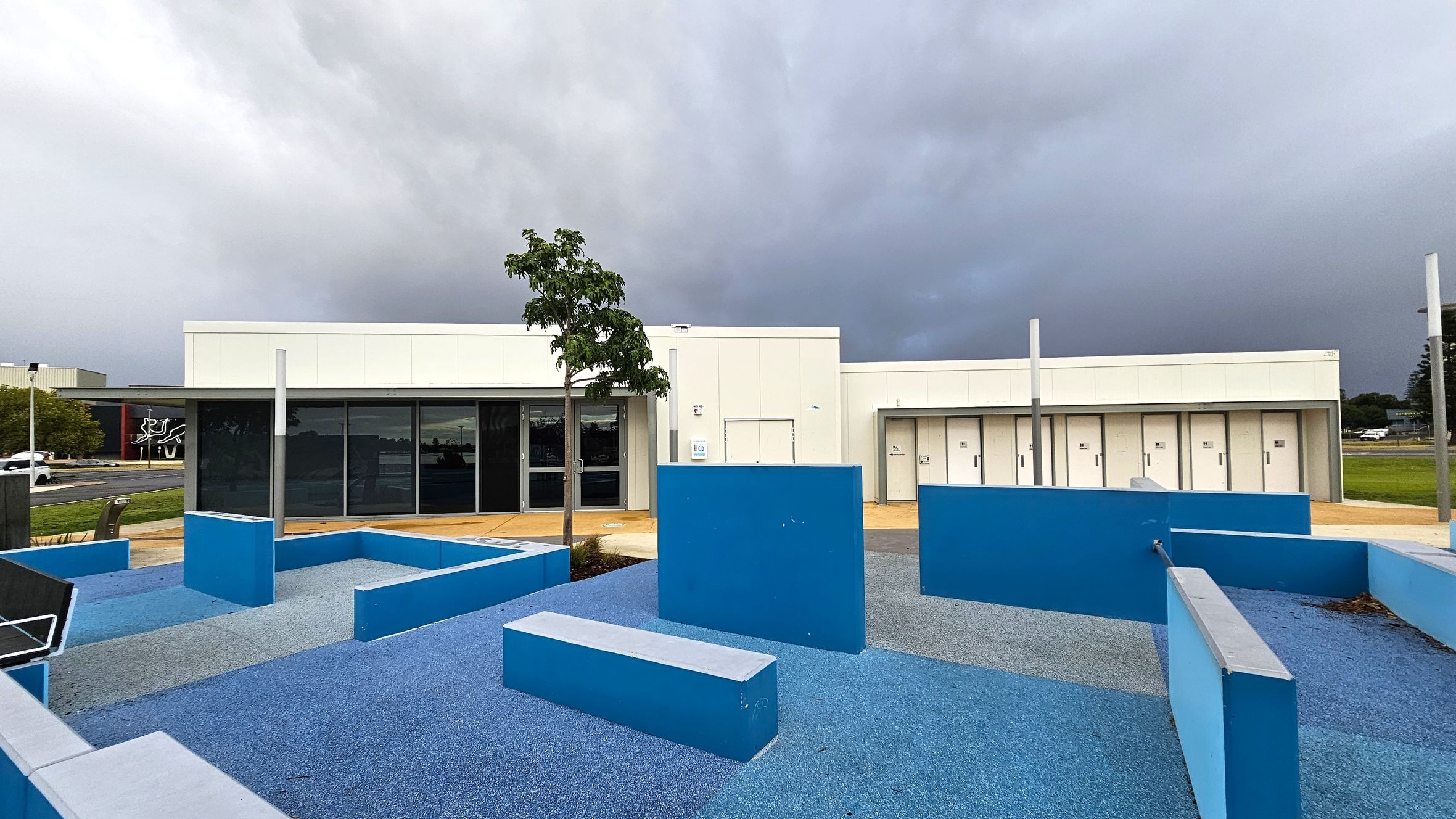 A modern building with a flat roof and white exterior is in the background. In front, there is a playground area with blue rubber flooring and various blue geometric structures, including benches and walls. A small tree is planted near the centre. To the left, a road with a few cars and a building with a mural can be seen. The sky is overcast, creating a grey backdrop.