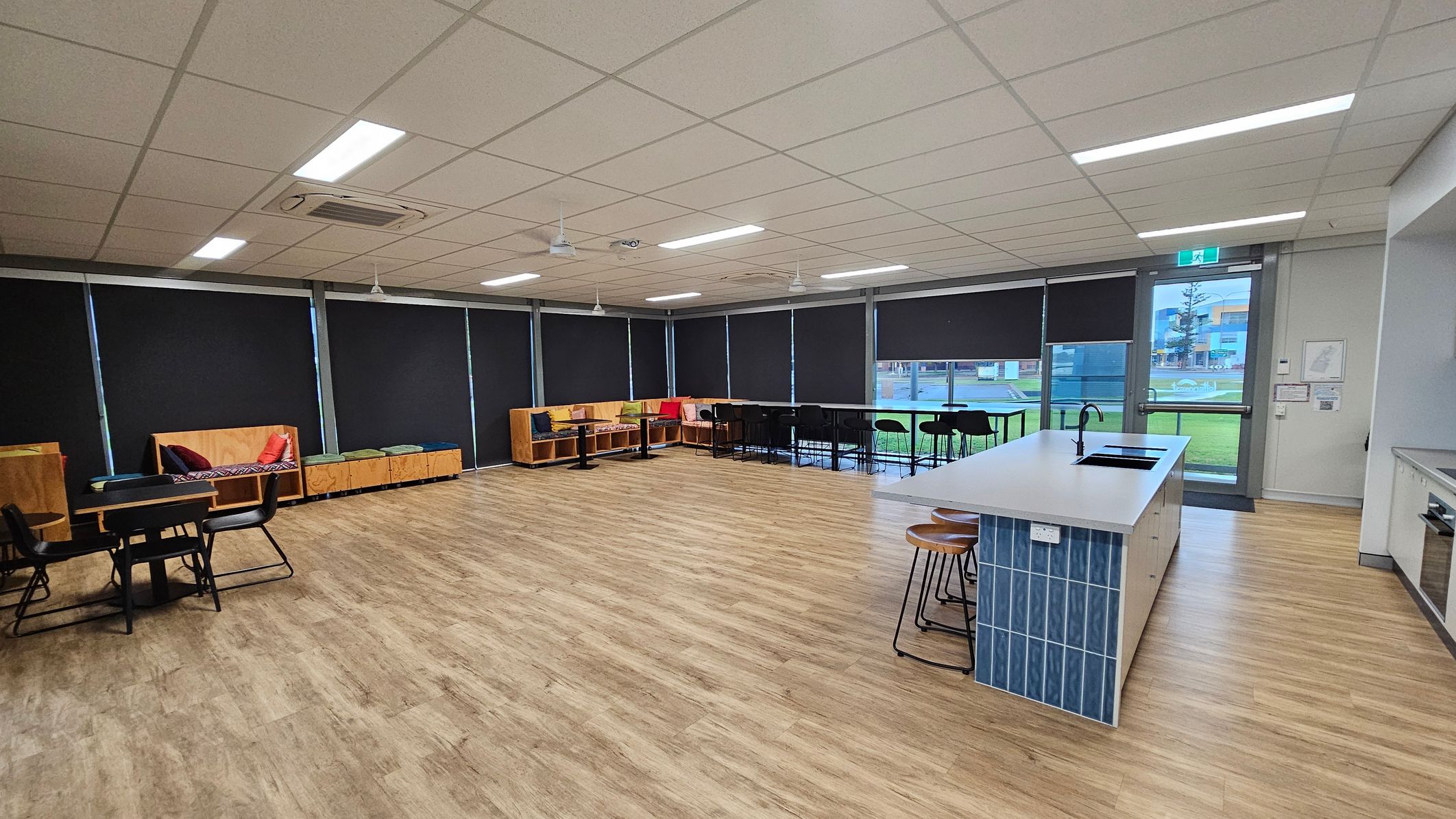 A spacious room with a light wooden floor and a white tiled ceiling. On the left, there are tables with black chairs and wooden benches with colourful cushions. The right side features a kitchen island with blue tiles and wooden stools. Large windows with black blinds line the back wall, and a glass door leads outside. The room is well-lit with fluorescent lights.