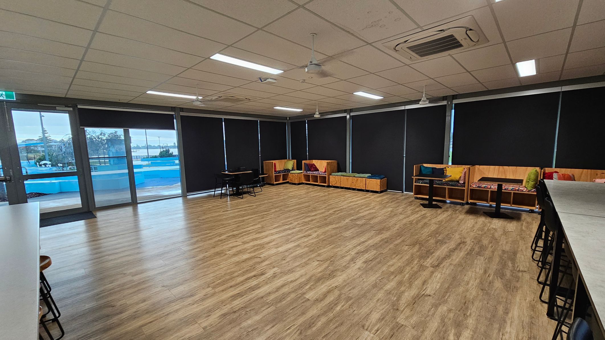 A spacious room with wooden flooring and a ceiling with fluorescent lights and air conditioning units. On the left, there are large windows with black blinds and a view of a blue outdoor area. In the centre, there are a few tables and chairs. On the right, there are wooden benches with colourful cushions. A long counter with high stools is also visible on the right side.