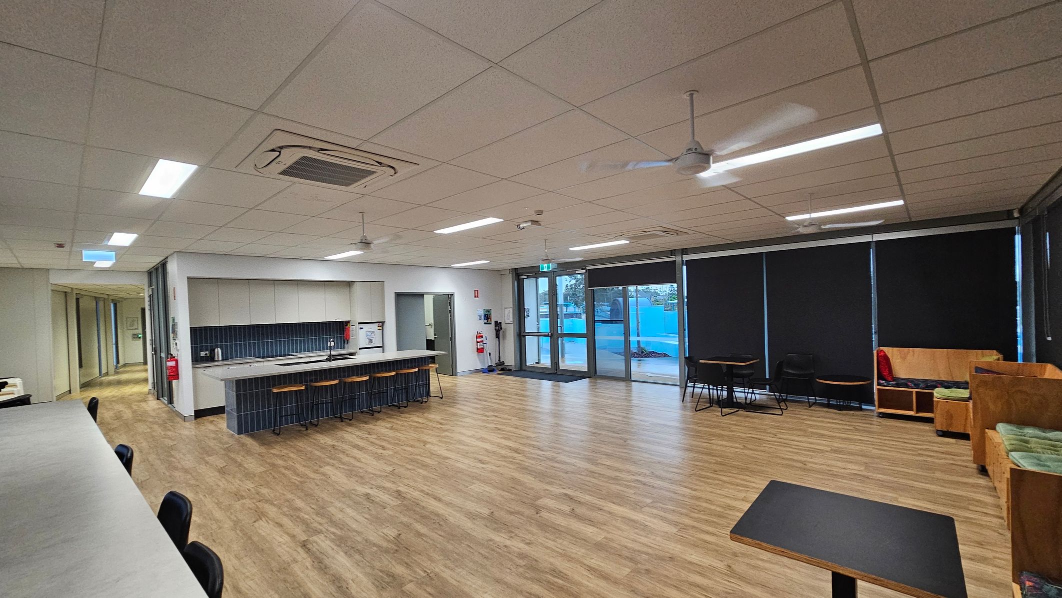 A spacious room with a light wooden floor and a tiled ceiling. On the left, there's a long counter with black chairs. In the middle, a kitchen area features a counter with stools, a sink, and cabinets. The right side has large windows with black blinds, a few tables, and wooden benches with cushions. Ceiling fans and lights are evenly spaced across the ceiling. A corridor is visible on the far left.
