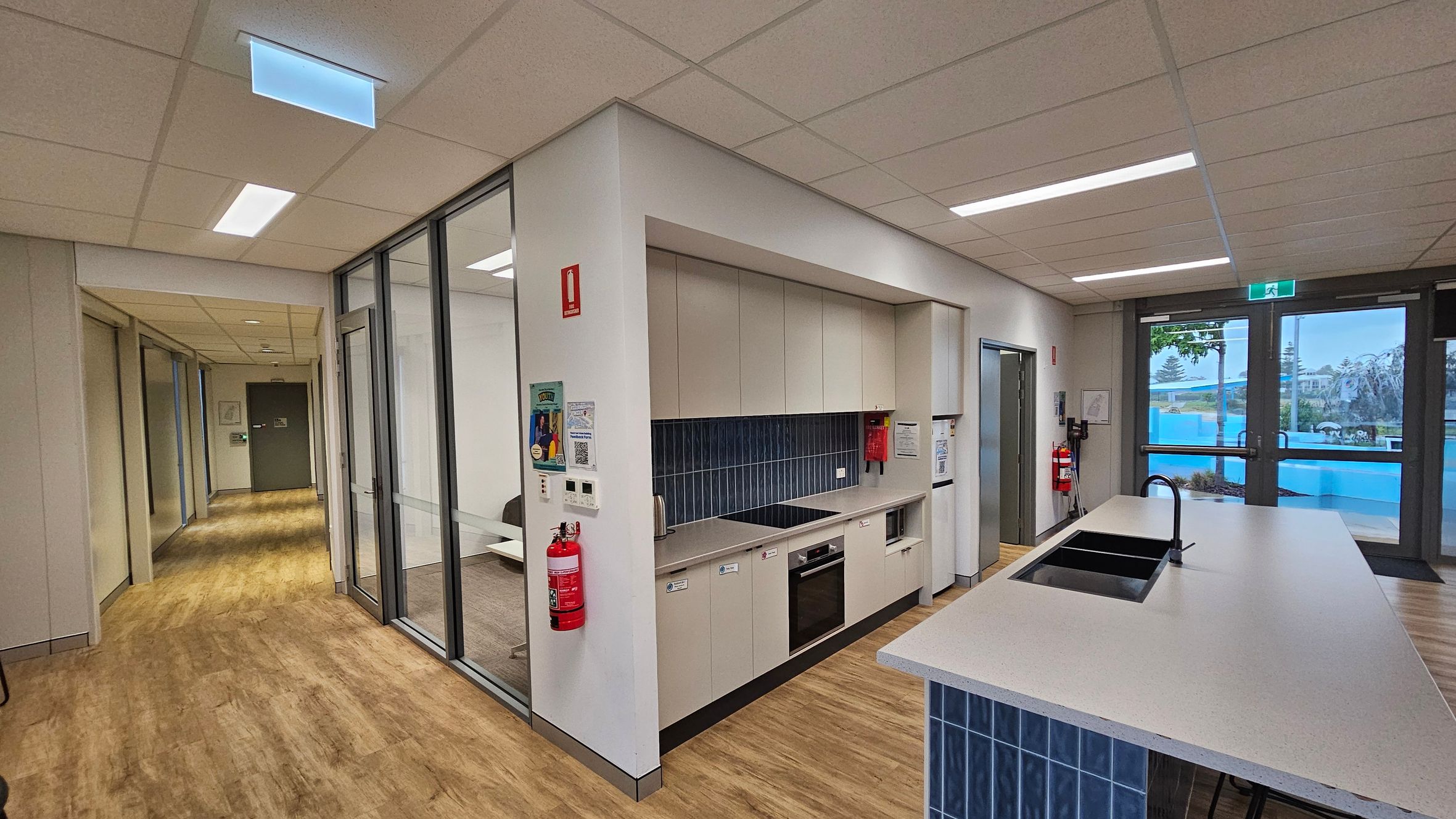 A modern kitchen area with light wood flooring and a suspended ceiling with recessed lighting. On the left, there is a hallway leading to a green door. In the centre, a kitchenette with beige cabinets, a black cooktop, and a built-in oven. A fire extinguisher is mounted on the wall. On the right, a large island with a sink and tap, and glass doors leading outside. Blue tiles are used as a backsplash and on the island base.