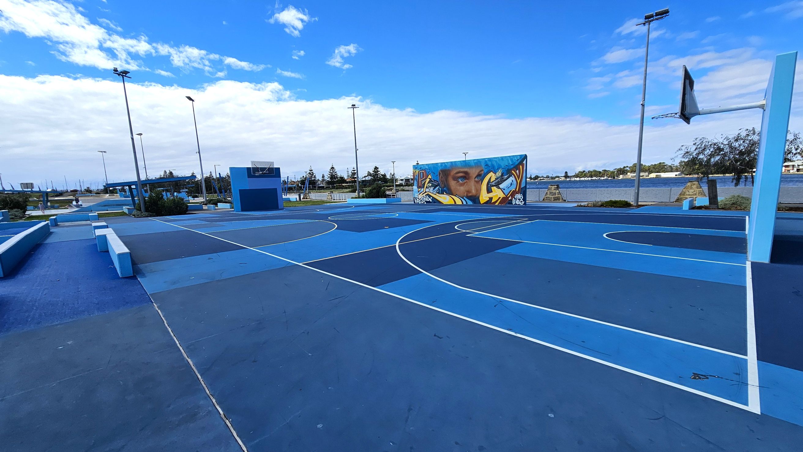 A vibrant outdoor basketball court with various shades of blue on the ground. On the right, a basketball hoop stands tall. In the background, a mural featuring a face and graffiti art is visible on a wall. Several tall streetlights are scattered around the area. Beyond the court, there is a body of water and some trees, with a few buildings in the distance. The court is surrounded by low benches and planters.