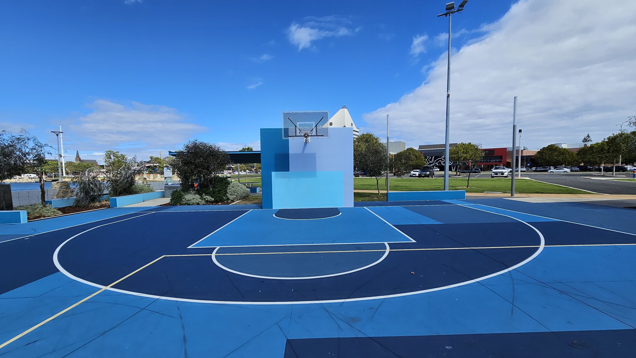 A vibrant outdoor basketball court with various shades of blue on the floor and backboard. The court features white lines marking the playing areas. In the background, there are trees and a grassy area, with a few parked cars and buildings visible. A tall streetlight stands on the right side. The sky is clear with some clouds.