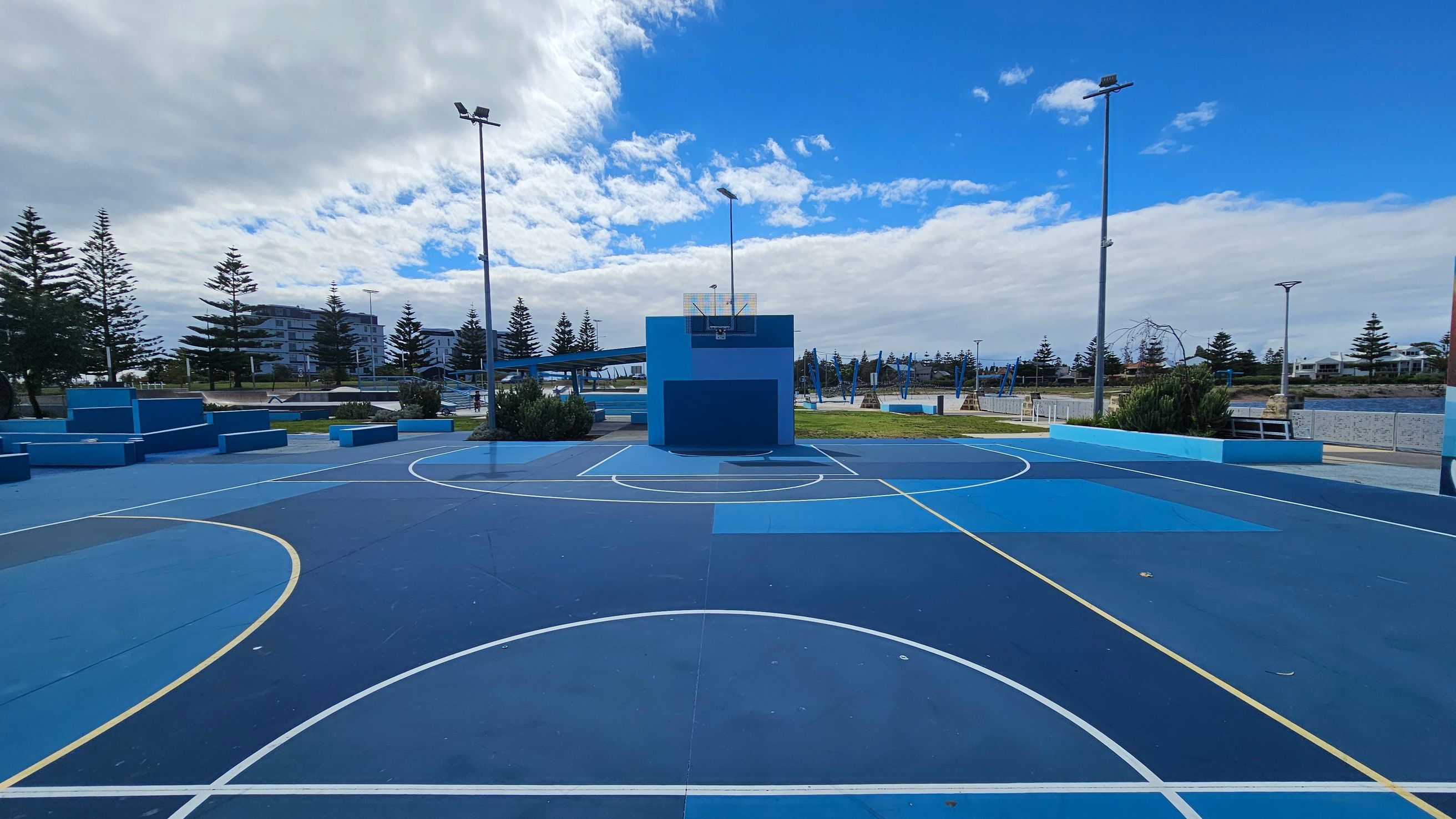 A blue outdoor basketball court with white and yellow lines. In the centre, a blue backboard and hoop are mounted on a blue wall. Surrounding the court are tall streetlights and some low bushes. In the background, there are several pine trees and modern buildings. The court surface is a mix of different shades of blue.