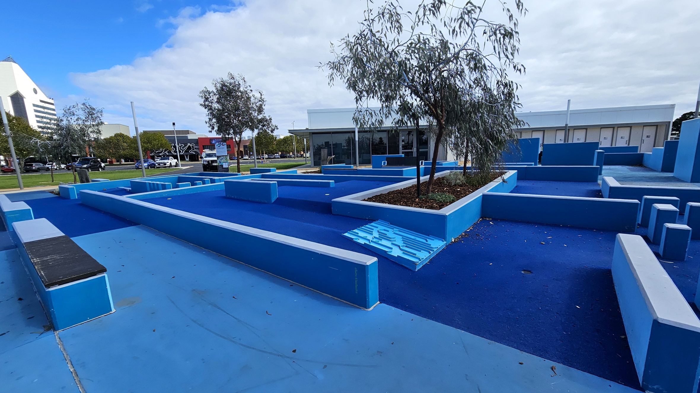 A parkour area with various blue platforms and obstacles of different heights and shapes. In the centre, there are small trees planted in raised beds. The ground is covered in blue rubberised material. In the background, there is a modern building with large windows. To the left, a street with cars and a building with a mural can be seen. The sky is partly cloudy.