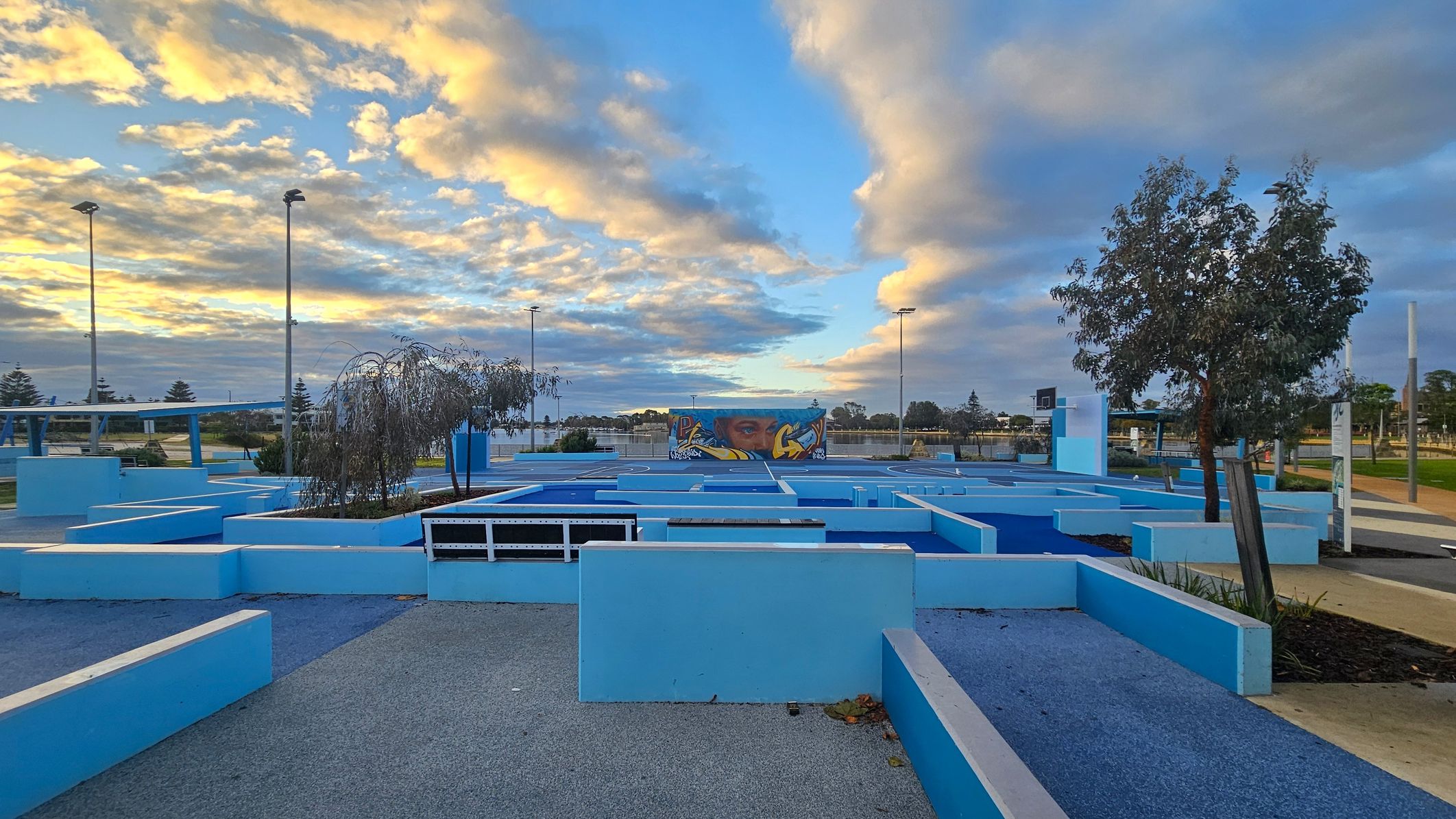 A modern outdoor space with geometric blue structures and pathways. In the centre, a mural with a face and graffiti art is visible on a wall. Trees are planted within some of the structures. Tall streetlights are scattered throughout the area. The ground is a mix of grey and blue surfaces, and there are patches of greenery. The sky is filled with clouds, illuminated by the setting or rising sun.