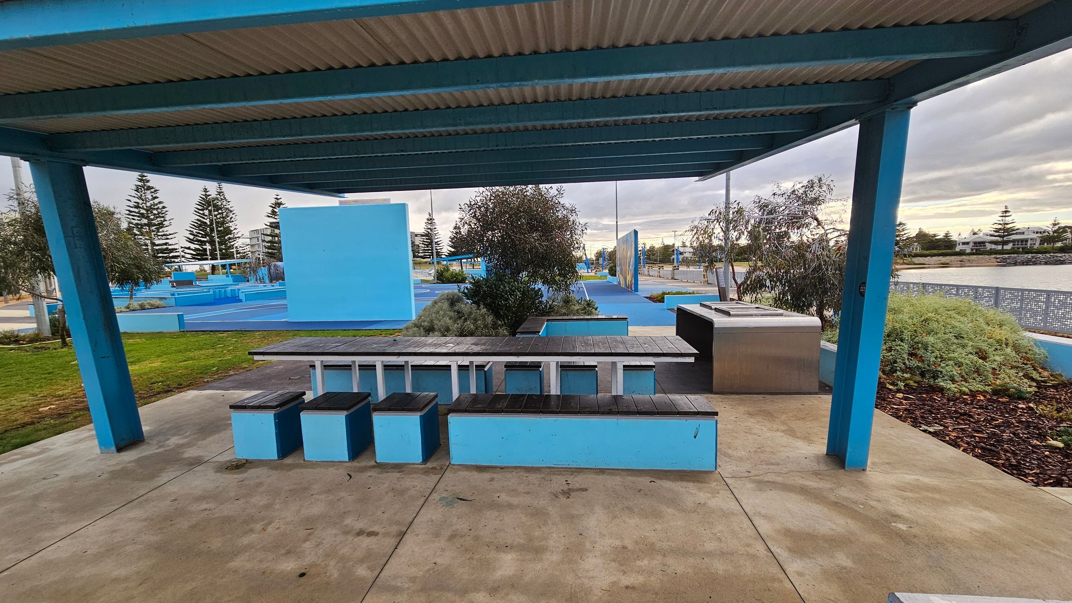 Under a blue metal shelter with a corrugated roof, there is a picnic area featuring a long table with attached benches. The benches are blue with black tops. To the right, a stainless steel barbecue is installed. The ground is a mix of concrete and grass. In the background, there are blue structures, trees, and a view of a body of water with more trees and buildings beyond.