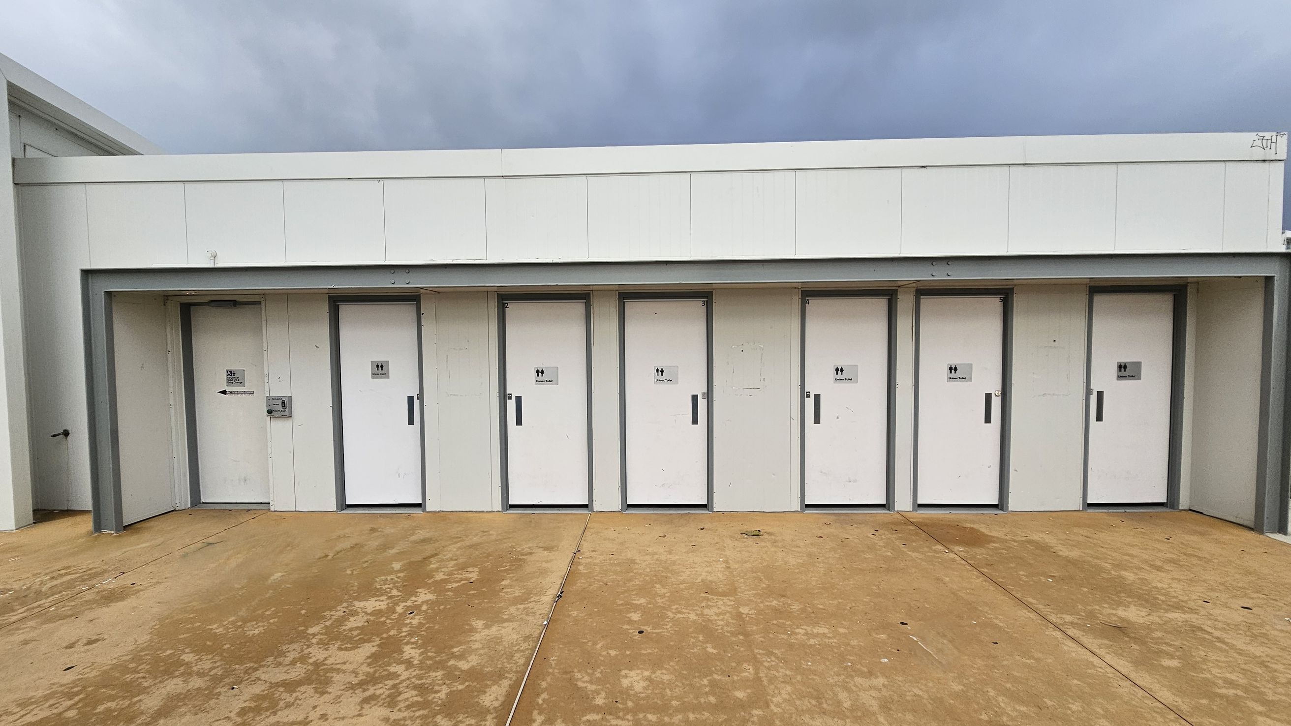 A row of six white doors, each with a unisex toilet sign, set into a white building. The doors are evenly spaced and have grey frames. The ground is a textured, light brown concrete surface. The building has a flat roof with a simple, modern design.