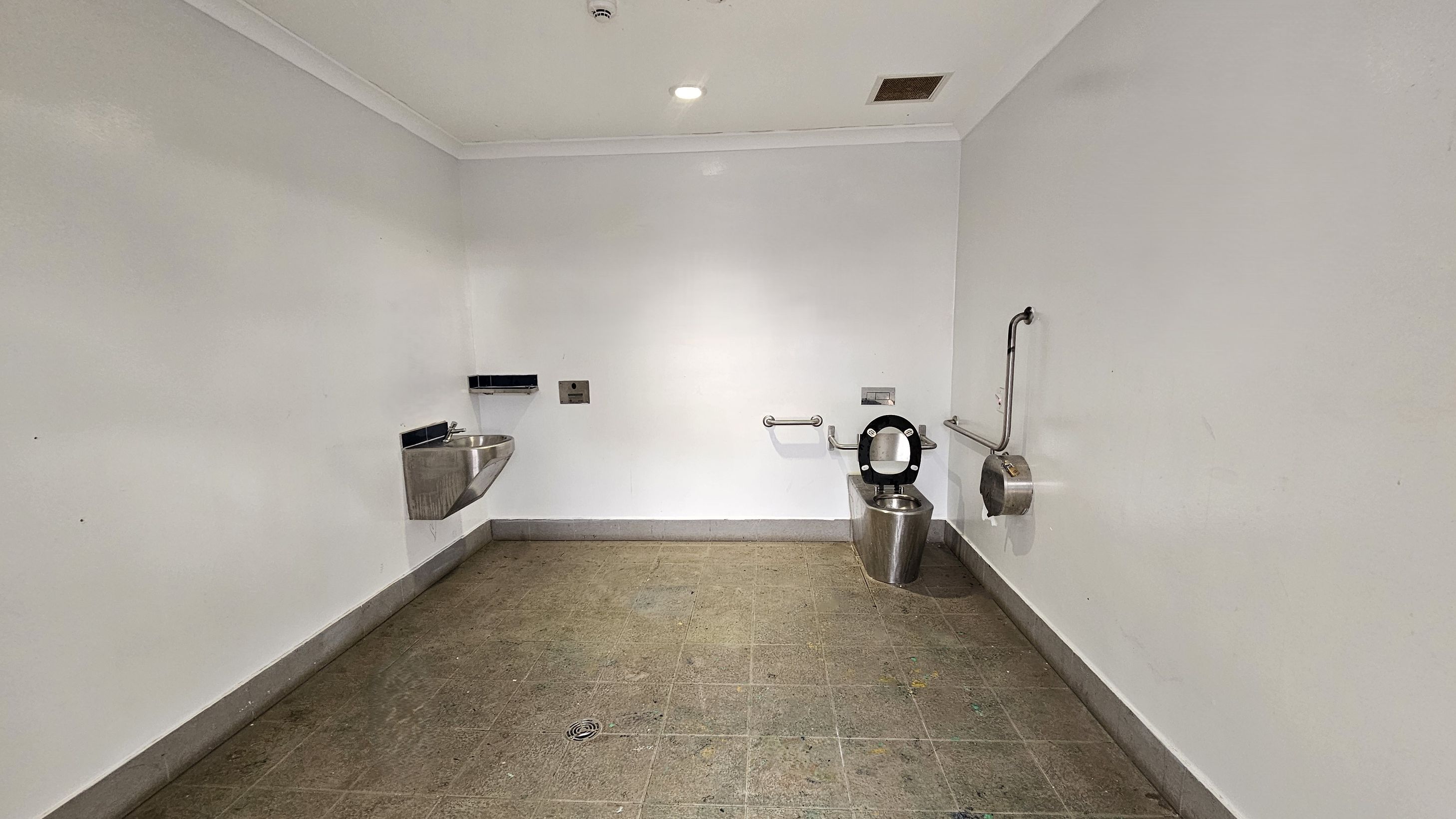 A public restroom with a stainless steel sink on the left wall, featuring a soap dispenser and a hand dryer above it. On the right, a stainless steel toilet with a black seat is installed, accompanied by grab bars for accessibility. A toilet paper dispenser is mounted nearby. The floor is tiled in brown with visible wear and stains. The ceiling is white with a recessed light and a vent. The walls are plain and painted white.