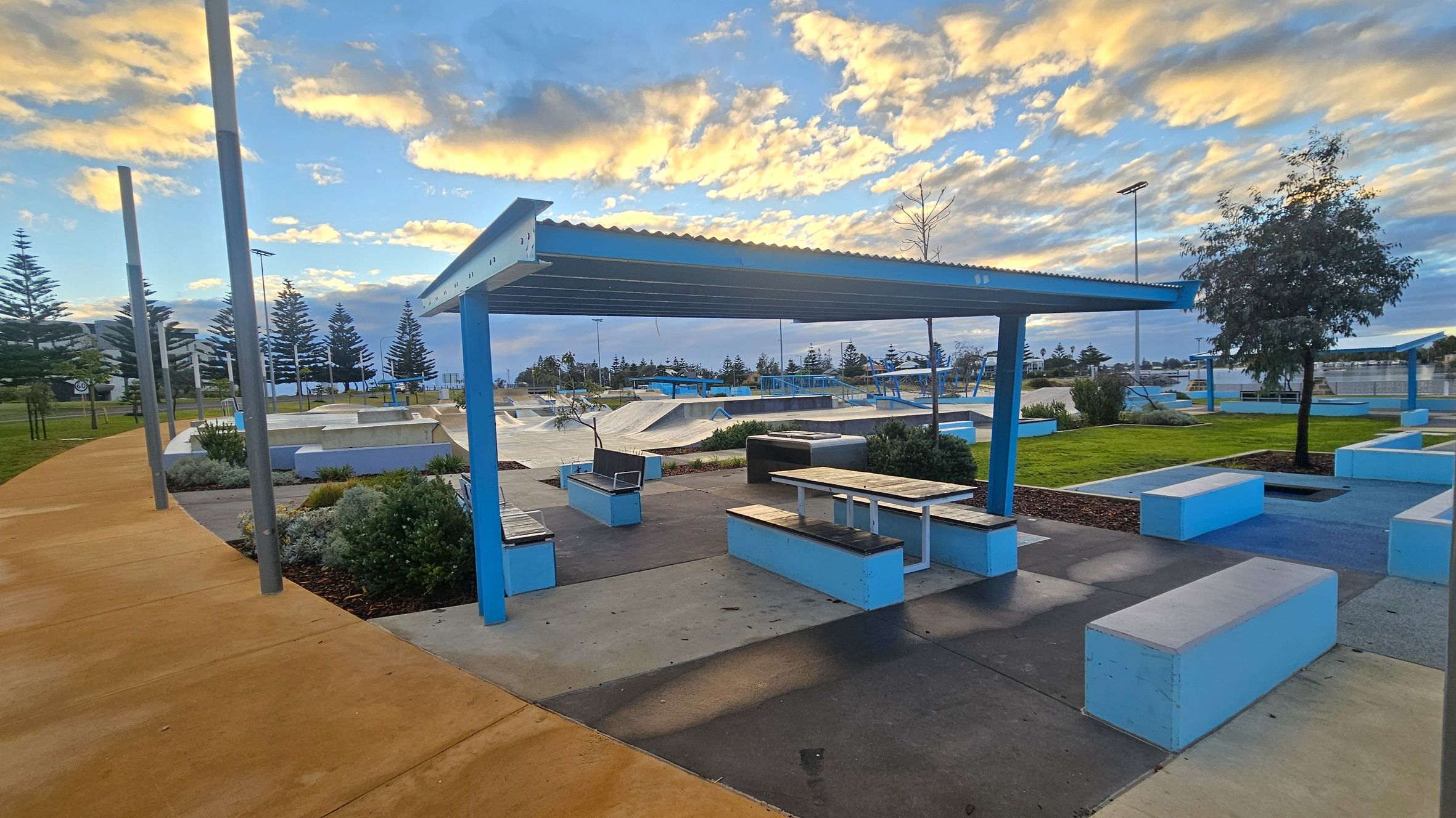 A skate park with a covered seating area in the foreground, featuring blue and white benches and tables. The ground is a mix of concrete and grass, with a yellow pathway on the left. In the background, there are skate ramps and more seating areas. Tall trees and lampposts are visible, with a line of pine trees further back. The sky is partly cloudy with a warm glow.